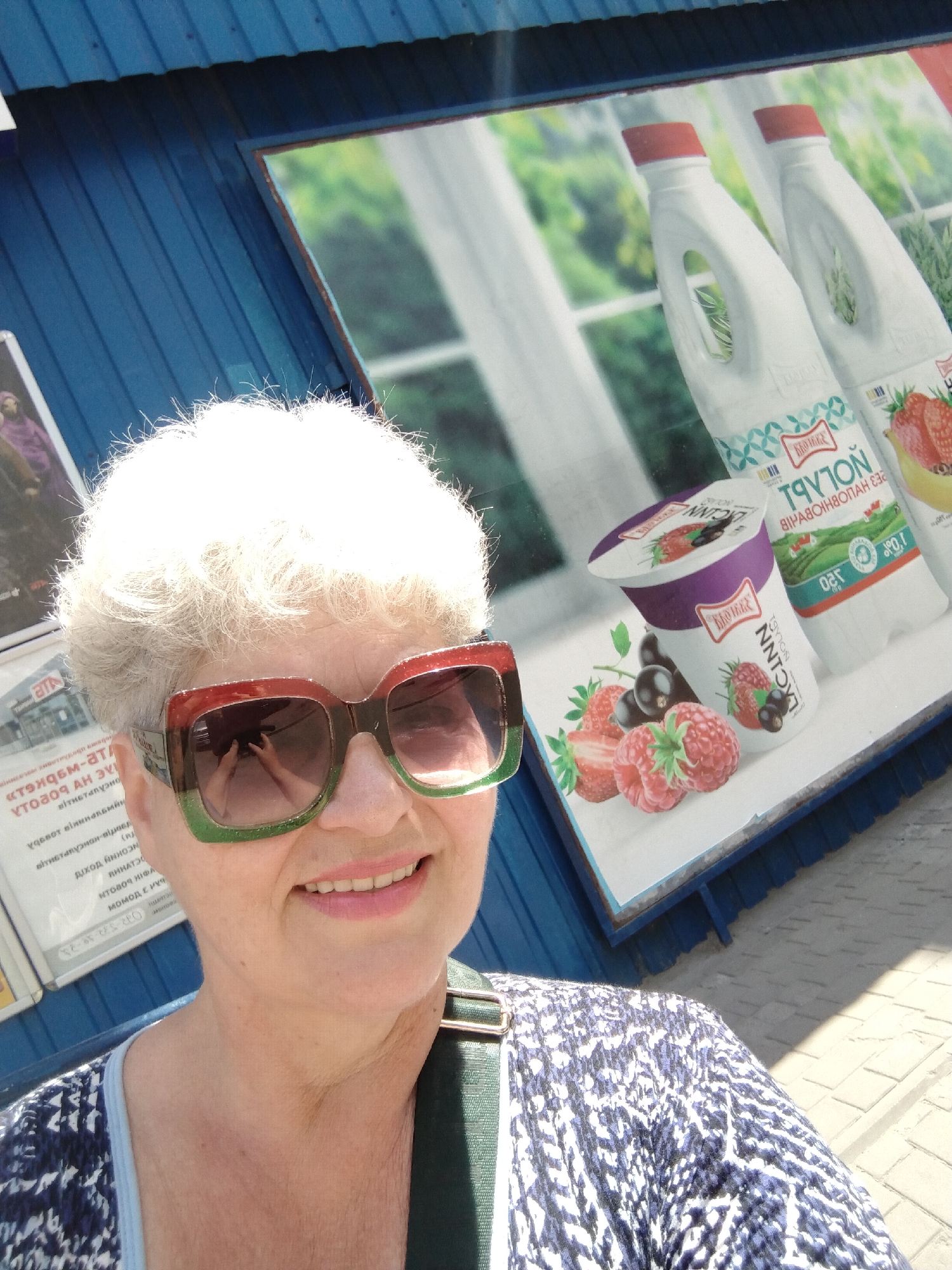 An eye-level, slightly high-angle, medium outdoor shot captures a white-haired woman with fair skin, wearing large square sunglasses with green and red frames and a blue and white patterned top with a green strap over her shoulder. She is smiling slightly and looking towards the camera. The background features a blue, corrugated metal structure and a large advertisement for dairy products, including bottles of milk and a yogurt cup with images of strawberries and berries. The advertisement is mounted on the blue structure, and the overall scene appears to be outdoors in a commercial area, possibly a street or market, in the city of Horishni Plavni, Ukraine.