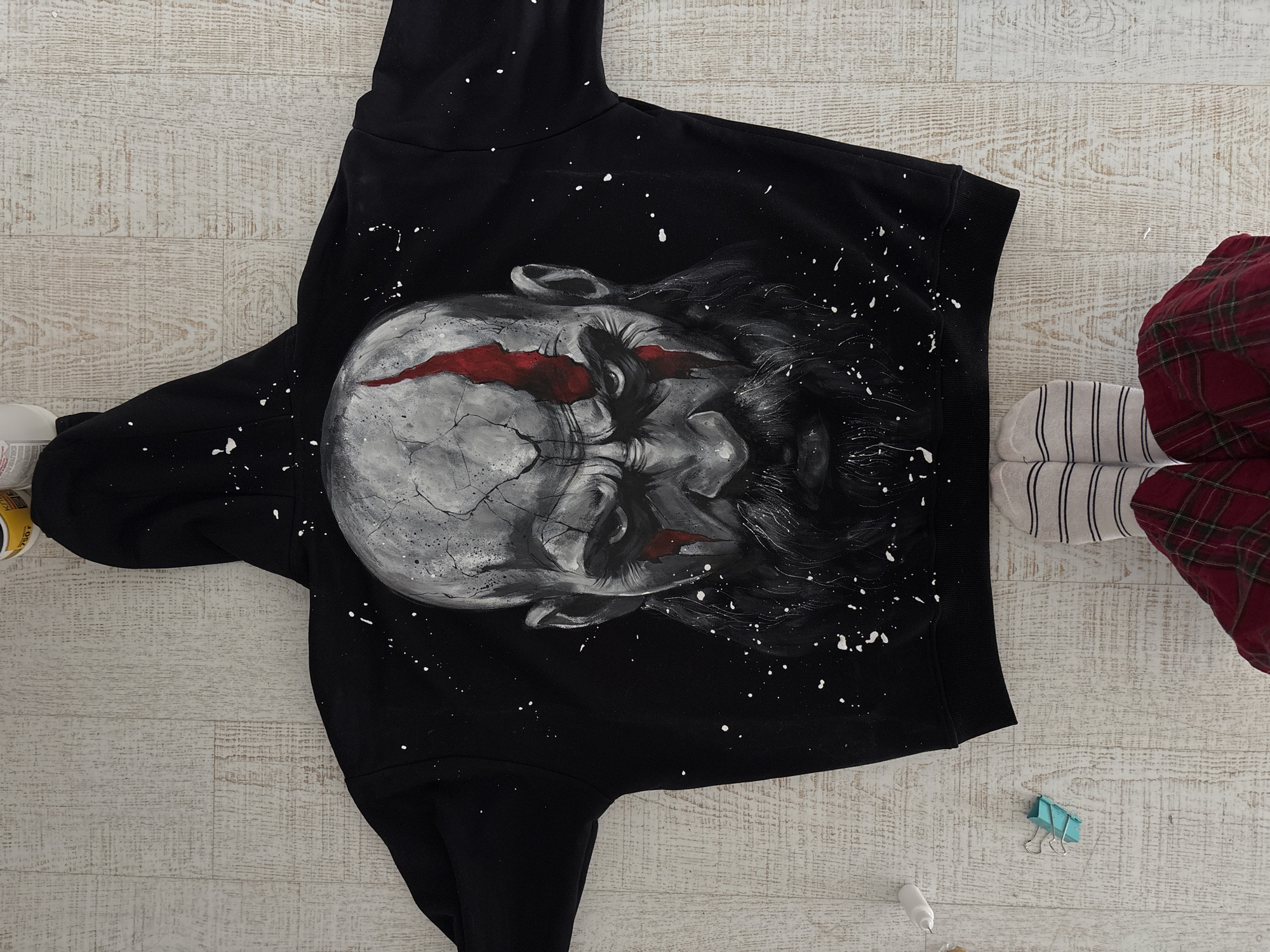 A black hoodie, custom-painted with a striking design, lies flat on a light-colored wooden floor. The central artwork on the hoodie is a stylized, somewhat abstract face or skull, rendered primarily in shades of grey and white. It features a textured, cracked appearance and intense red marks around the eyes and forehead, with dark, flowing elements resembling hair extending outwards. Numerous white paint splatters are scattered across the black fabric, creating a starry or distressed effect.
To the right of the hoodie, part of a person's lower legs and feet are visible. They are wearing red plaid trousers and white socks with thin black horizontal stripes, suggesting a casual, indoor setting.
Several small objects are scattered around the scene. A light blue binder clip rests on the wooden floor near the person's feet. A small white bottle or tube, likely containing paint, is partially visible at the bottom right corner. On the far left, a yellow container with "K 750" and further illegible text is partially visible, indicating art supplies.
The wooden floor consists of light-toned planks, providing a clean and bright background. The arrangement of the items and the presence of paint supplies suggest an ongoing artistic or crafting activity. The setting appears to be an indoor space, possibly a home or studio in Krasnoyarsk, Russia.