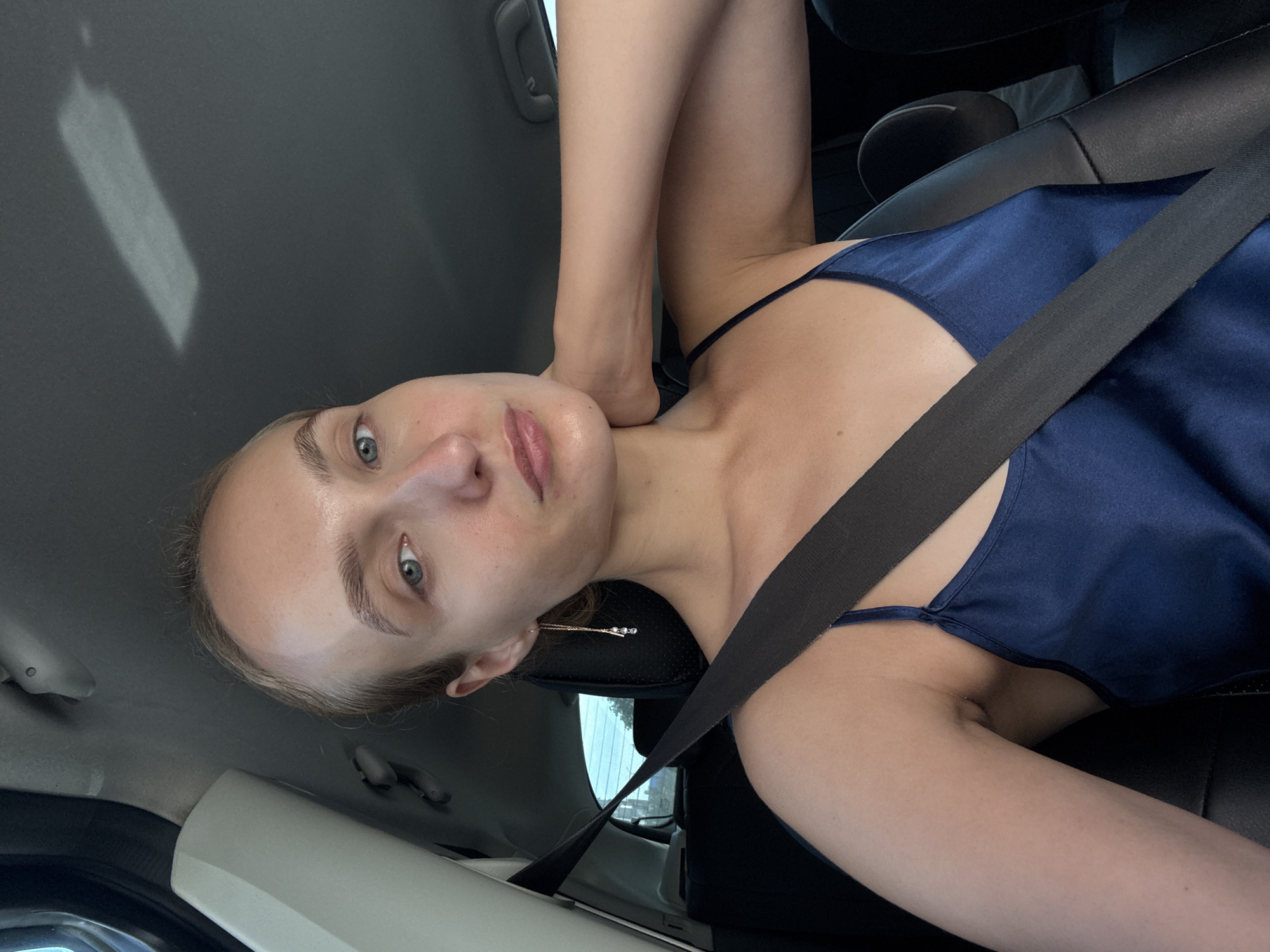 A young woman with fair skin and light hair is sitting in the driver's seat of a car. She is wearing a dark blue satin slip dress and a black seatbelt. She has blue eyes and light eyebrows. The interior of the car is visible, including the dashboard, steering wheel, and seats. The car appears to be parked or moving slowly. The lighting suggests it is daytime. The specific location within Votkinsk, Russia, cannot be determined from the image alone.