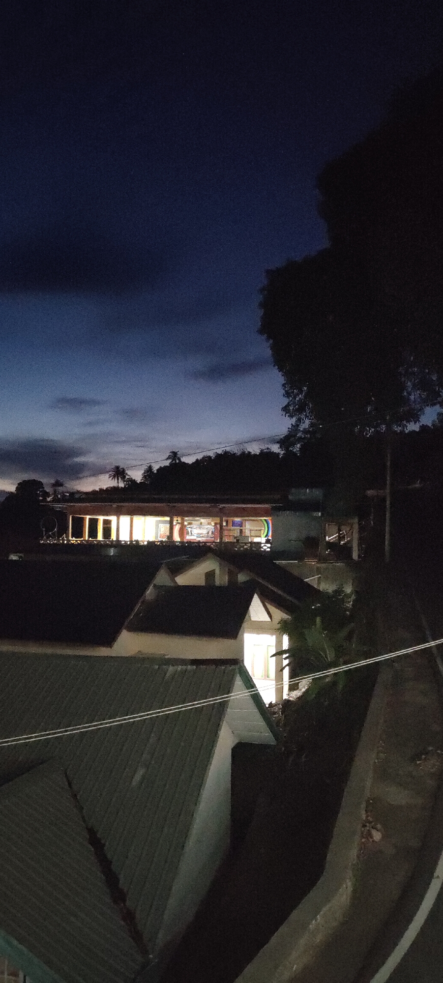 The image shows a view of buildings at dusk in Sabang, Indonesia. In the foreground, there are several pitched roofs of residential buildings, some with green corrugated metal roofing. In the mid-ground, a longer, rectangular building is illuminated from within, suggesting it is a commercial establishment or community center. This building features large windows and appears to have a covered walkway or veranda along its front. In the background, a dark, tree-covered hillside rises, with a few palm trees visible. The sky is a deep blue, transitioning from lighter hues near the horizon, indicating either early morning or late evening. The overall scene depicts a quiet, residential and commercial area in Sabang during twilight.