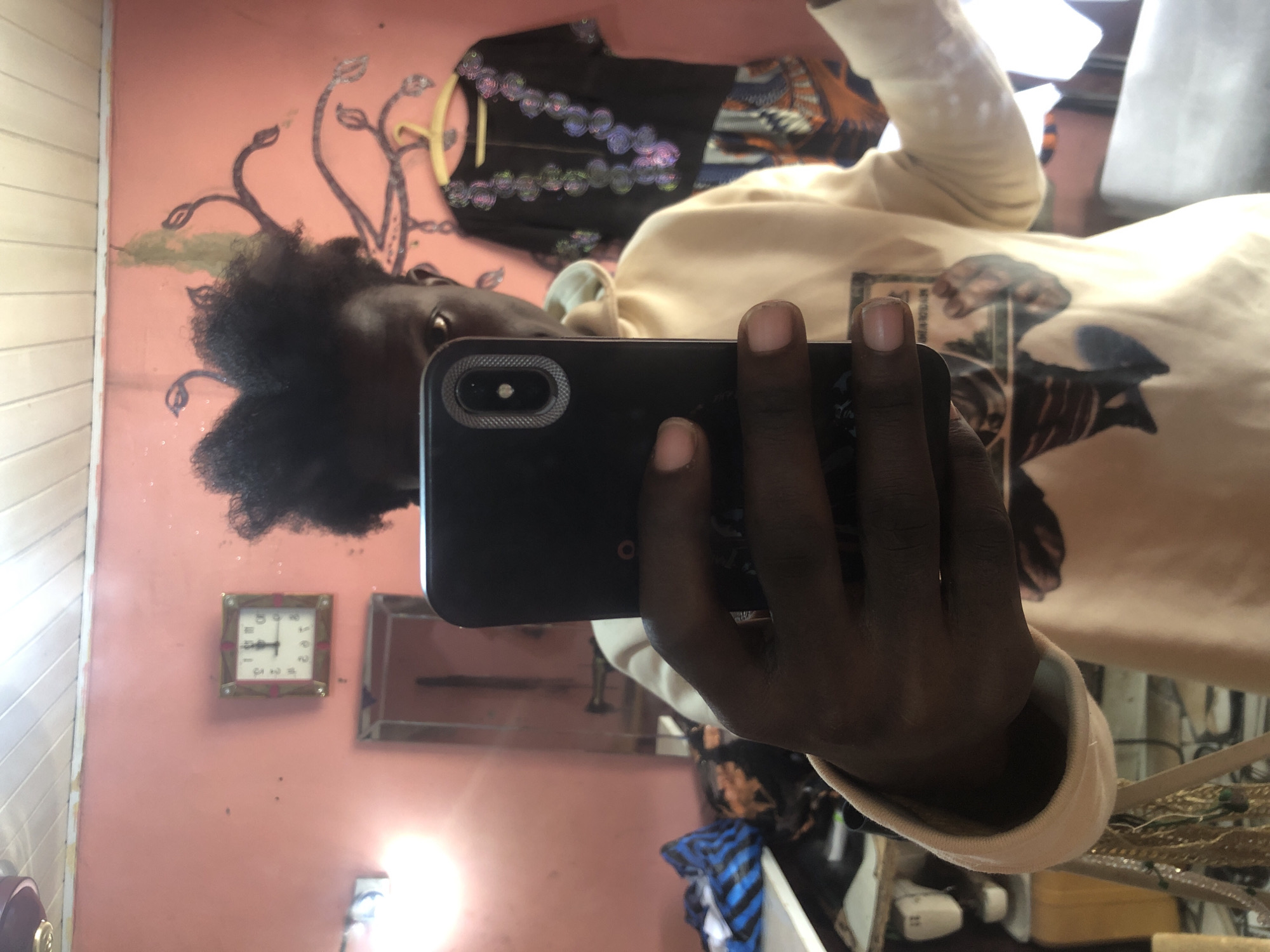The image is a selfie taken in a room in Ado, Nigeria. A dark-skinned person with curly hair is holding a smartphone, taking the picture in a mirror. The person is wearing a light-colored hoodie. Behind them, a pink wall is visible, adorned with a square clock showing the time as approximately 1:05. A decorative vine motif is also painted on the wall. To the right, a black garment with colorful embroidery hangs on a hanger. In the background, a blurred reflection shows furniture and other items in the room.
