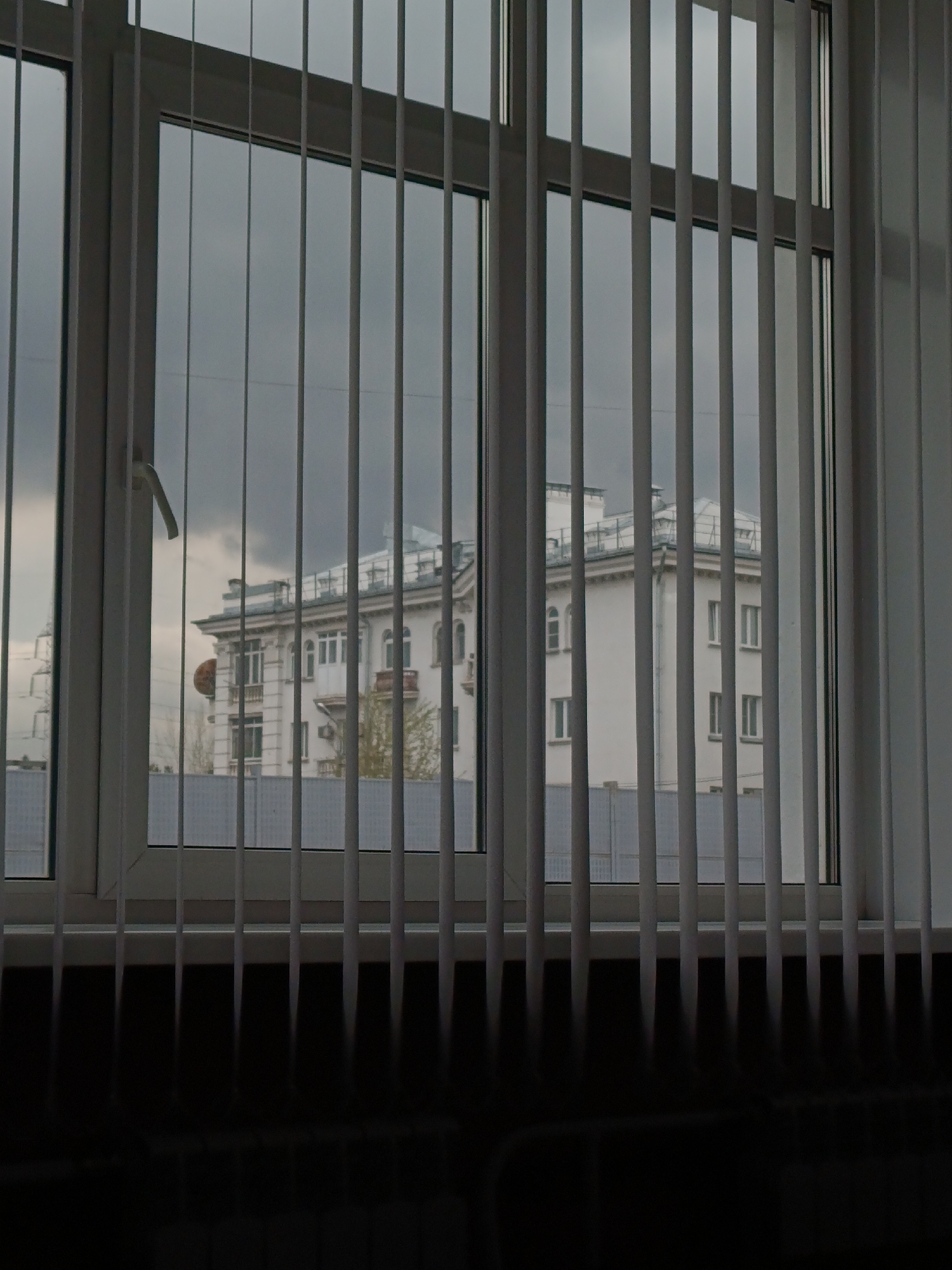 A building with white walls is seen through a white-framed window with vertical blinds. The sky is overcast, and there is a fence visible in the background. The location is suggested to be Krasnoyarsk, Russia.