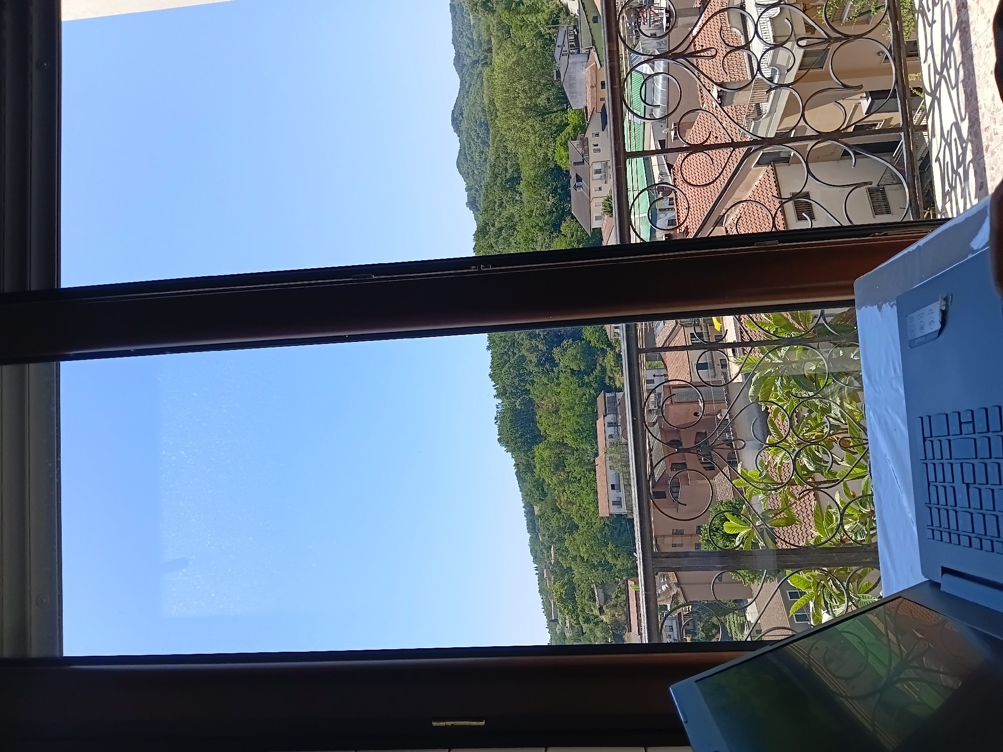 The image depicts a view from a window overlooking a town in Vallesaccarda, Italy. In the foreground, a laptop is placed on a white surface, partially obscuring the view. The window frames a scene of buildings with red-tiled roofs, some with balconies and ornate iron railings. Beyond the town, lush green hills covered in trees rise up, with a clear blue sky above. The overall impression is one of a serene, sun-drenched day.