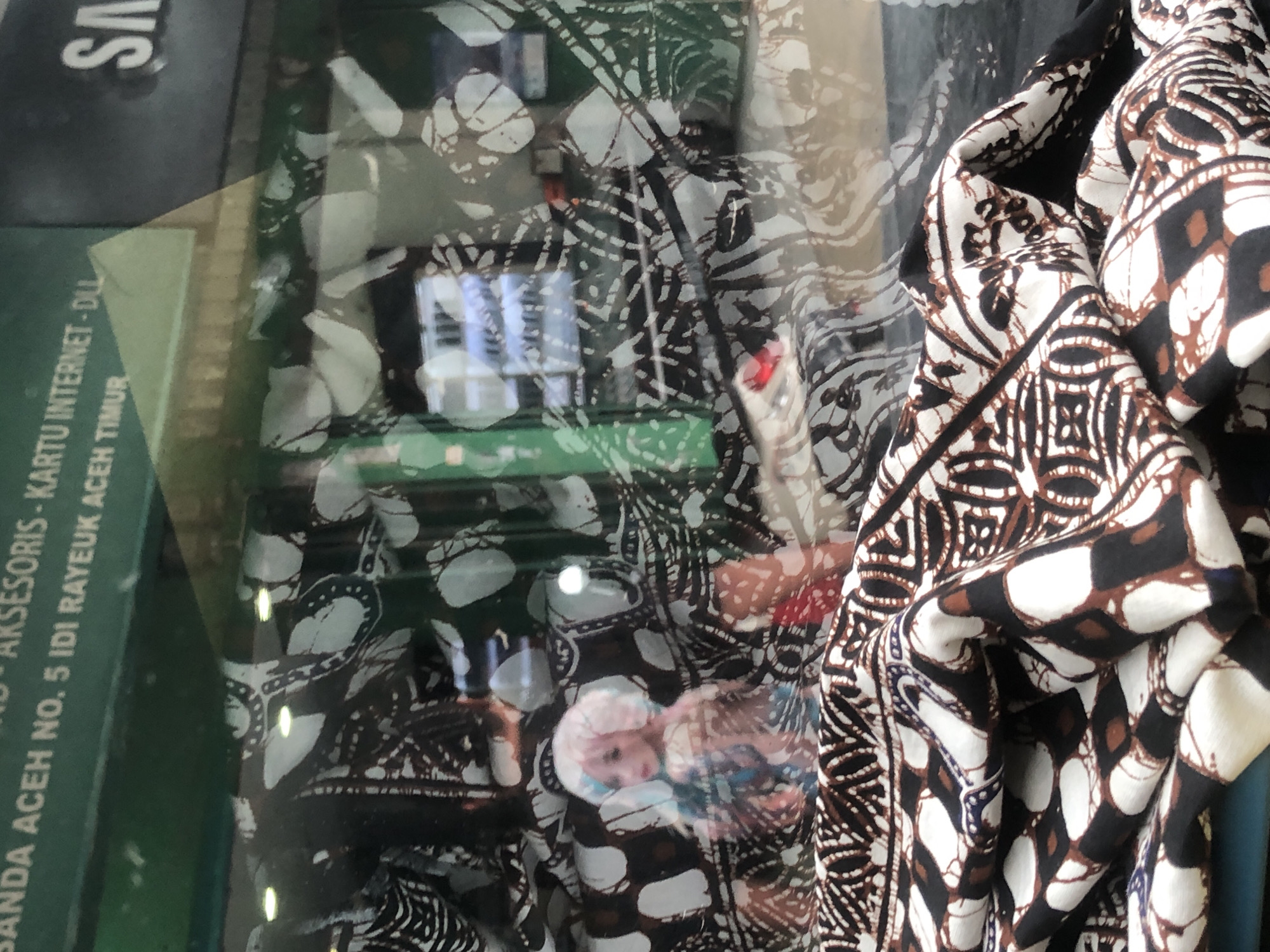 The image shows a store window displaying merchandise. In the foreground, a richly patterned textile, likely batik, is folded and draped. Behind the textile, a doll with pink and blue hair and a light-colored dress is visible. The doll is positioned in front of a wall adorned with a black and white patterned fabric. Reflections in the glass indicate an outdoor environment with a sign that reads "ANDA ACEH NO. 5 IDI RAYEUHK ACEH TIMUR" and another sign with the "SAMSUNG" logo. The overall impression is of a retail shop in Titibaro, Indonesia, showcasing textiles and possibly other items.