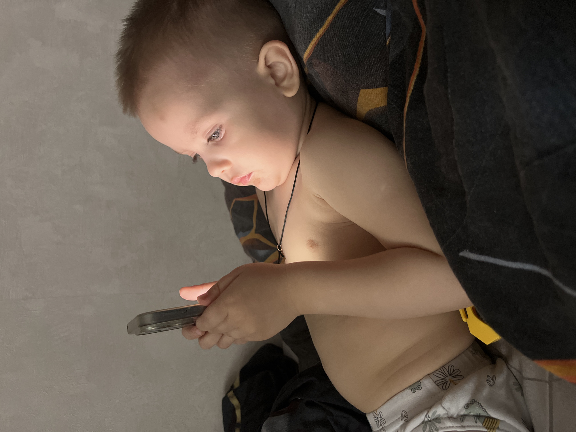 A young child is lying in bed, seemingly in a home, illuminated by the light from a cell phone they are holding in their hands. The child is bare-chested, wearing patterned shorts. A necklace is visible around their neck. Part of a dark-colored blanket is draped over the child. The background consists of a light-colored wall. There is no explicit information in the image itself to indicate a location in the city of Марковский, Russia, therefore, that information cannot be confirmed.