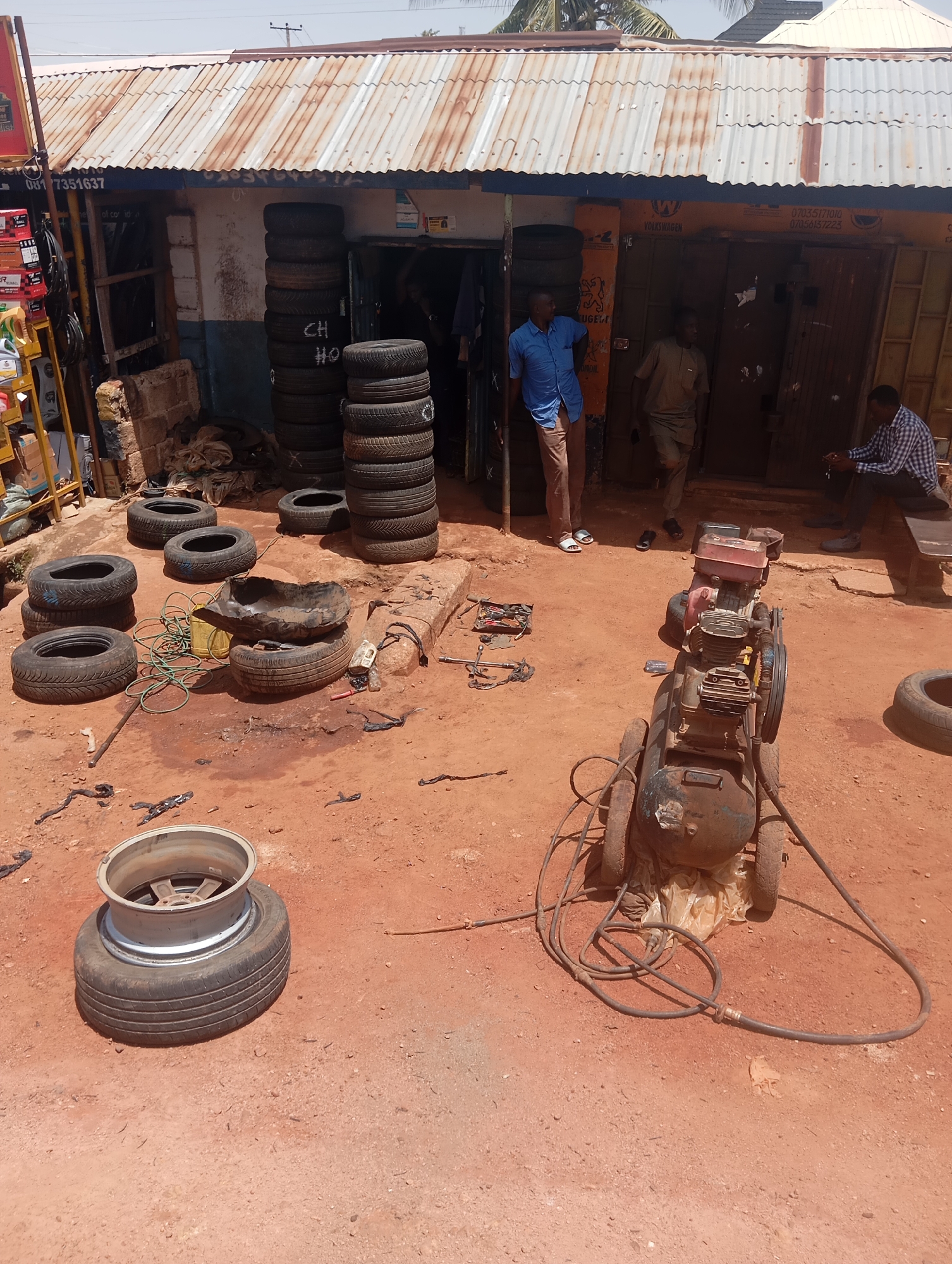This is an outdoor scene in Jos, Nigeria, depicting a tire repair shop. In the foreground, a stack of tires is visible, with one tire and rim assembly prominently displayed. To the right, an old, rusty air compressor with hoses sits on the dirt ground. Scattered around the compressor and tires are various tools, including a wrench and other metal objects, as well as some plastic sheeting and a yellow bucket. In the background, a building with a corrugated metal roof houses the tire shop. Several individuals are present: a man in a blue shirt stands near the entrance, another man in a beige outfit walks into the shop, and a third man in a checkered shirt sits on a bench outside. The environment is dusty, with red earth covering the ground.
