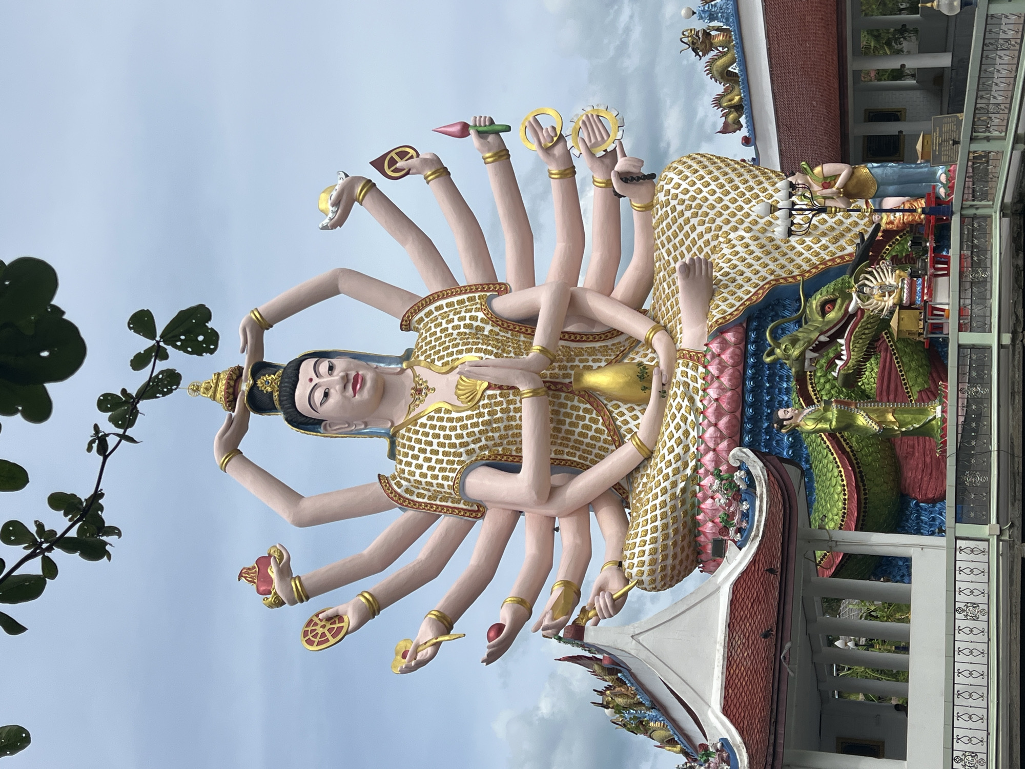 An outdoor, eye-level, full shot captures a large, multi-armed statue in Baan Plai Laem, Thailand, against a cloudy sky with green foliage framing the top. The statue, adorned in white and gold, sits cross-legged with hands in a prayer pose. Multiple arms extend from its torso, each holding a symbolic object, including a lotus flower, a wheel, a red orb, and a flame. Below the statue, a vibrant green dragon sculpture winds around its base. Two small human figures, one in green and one in yellow and blue, stand beside the dragon. In the background, traditional Thai temple architecture with red roofs and decorative elements is visible. The scene is serene, with no discernable action or interaction.