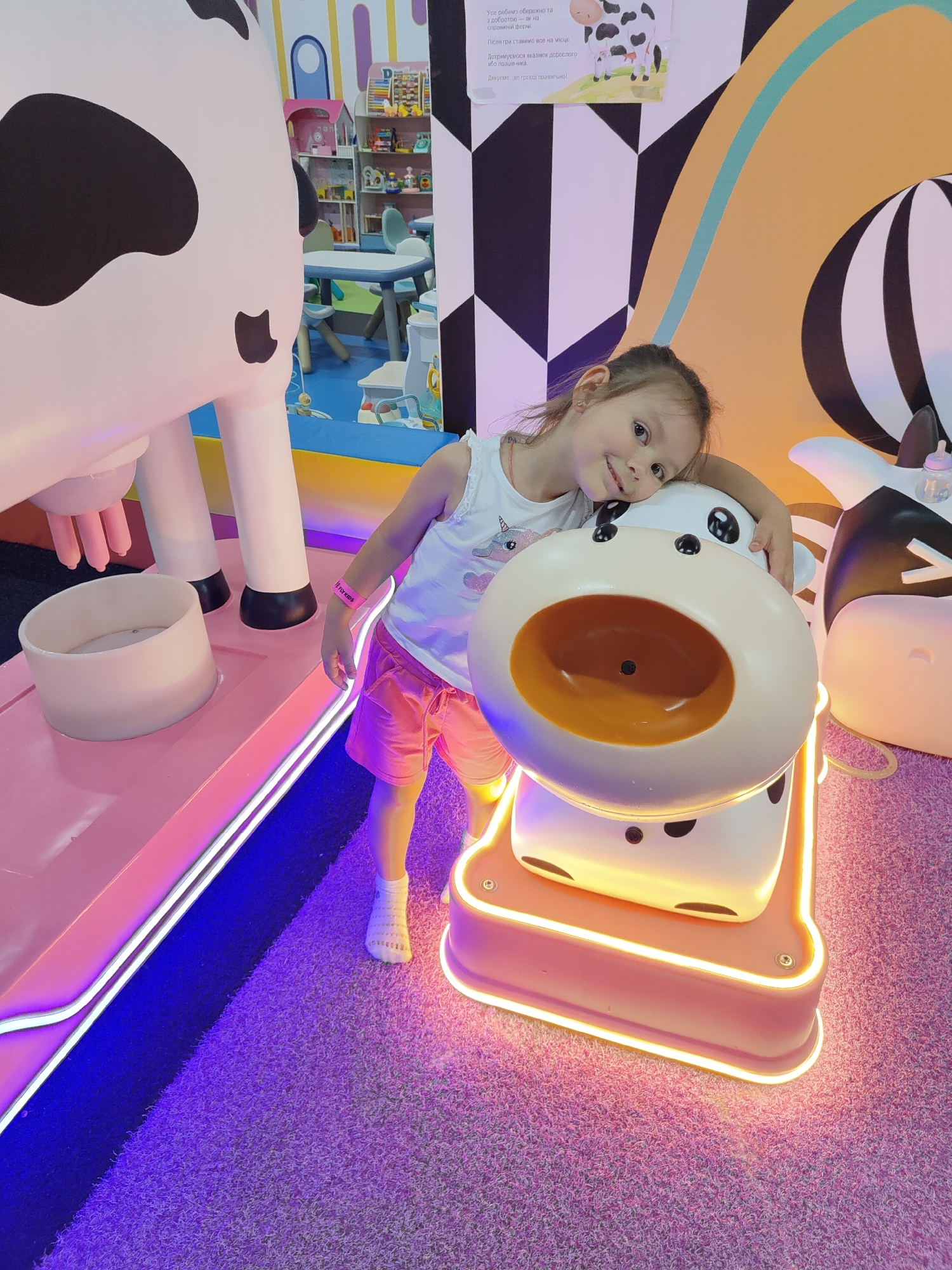 This is a medium shot, eye-level photograph of a young girl in an indoor playground. The girl is leaning against a large, cartoonish cow-themed structure. She is smiling and looking directly at the camera, with her head resting on the cow's head. She is wearing a white tank top with a unicorn print, pink shorts, and white socks. On her wrist is a pink wristband with "FLY KIDS" printed in white letters.
The cow structure appears to be part of a play area, possibly a feeding or interactive station for children. It is predominantly white with black spots and has a large, open mouth area that is a light brown color. The cow is mounted on a pink platform with neon lighting around its base. Behind the girl, another, larger cow structure is partially visible on the left.
The background of the playground features colorful, geometric patterns on the wall in black, white, and orange. To the left, a glimpse of a play area with shelves filled with toys and small tables and chairs suggests a dedicated children's entertainment space. The floor is covered in a textured pink carpet. The lighting is bright and artificial, creating a vibrant and playful atmosphere. The overall mood is cheerful and engaging, as conveyed by the girl's happy expression and the colorful, whimsical surroundings. The text in the image is in Ukrainian and appears to be instructions or a thank you message for playing correctly. The location is identified as Kryvyi Rih, Ukraine.