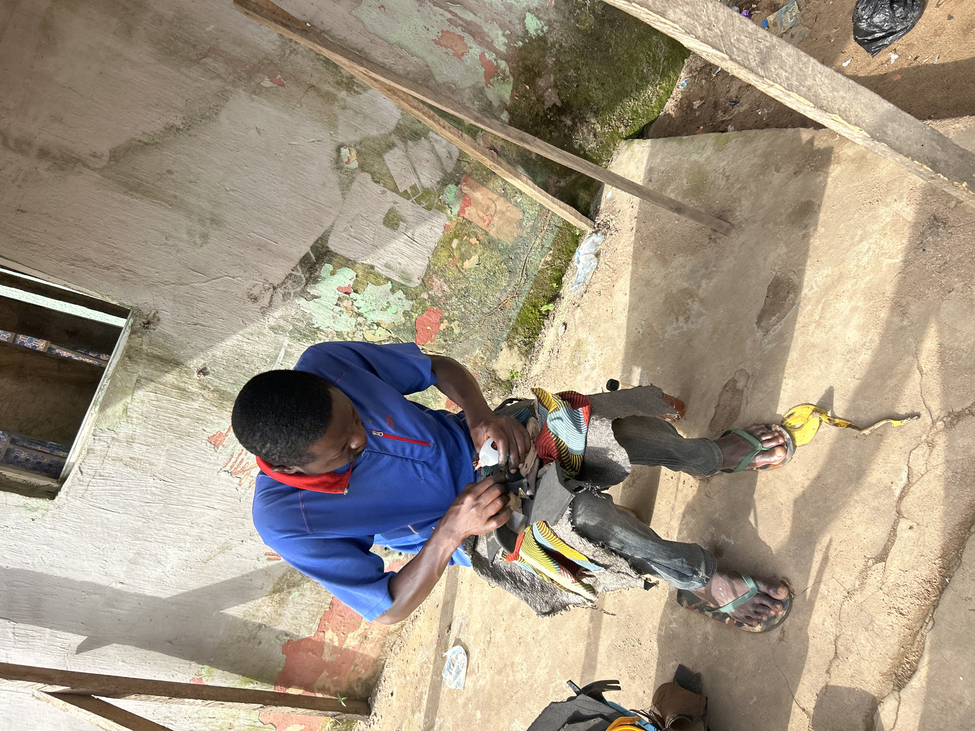 An outdoor scene in Ihiagwa, Nigeria shows a man sitting on a low stool and working on what appears to be shoes. He is wearing a blue t-shirt with a red collar and grey pants. The man is holding a white object, possibly glue or sealant, in his right hand and is manipulating dark-colored shoe material with his left hand and tools. The stool he is sitting on is covered with a patterned cloth, and he is wearing flip-flops. In front of him, on the ground, is a discarded banana peel. The background consists of a rough, weathered concrete wall with peeling paint in green, red, and white, and some moss growth. Two wooden poles lean against the wall. The ground is dirt and concrete.