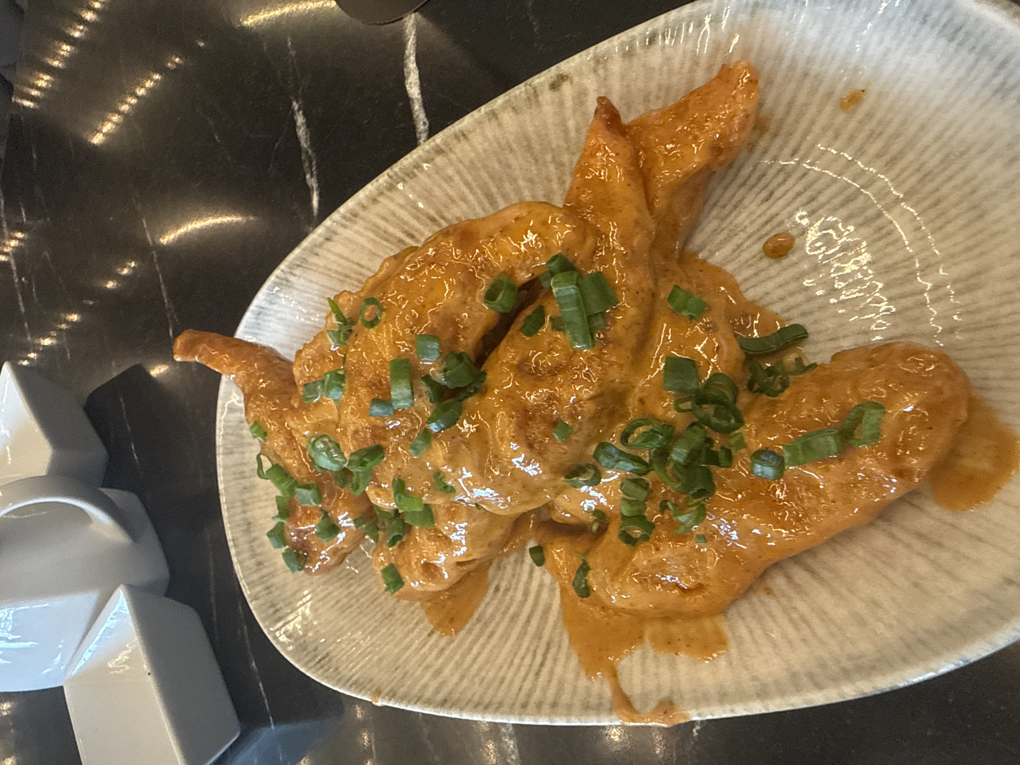 A high-angle, indoor shot depicts a plate of fried chicken wings with a creamy orange sauce, garnished with chopped green onions. The wings are arranged on a shallow, circular plate with a textured, gray and white pattern. The plate sits on a dark, reflective surface, possibly a table with a marble or polished stone finish. To the left of the plate is a stack of white, geometric serving dishes. The photograph appears to have been taken in Puebla, Mexico.