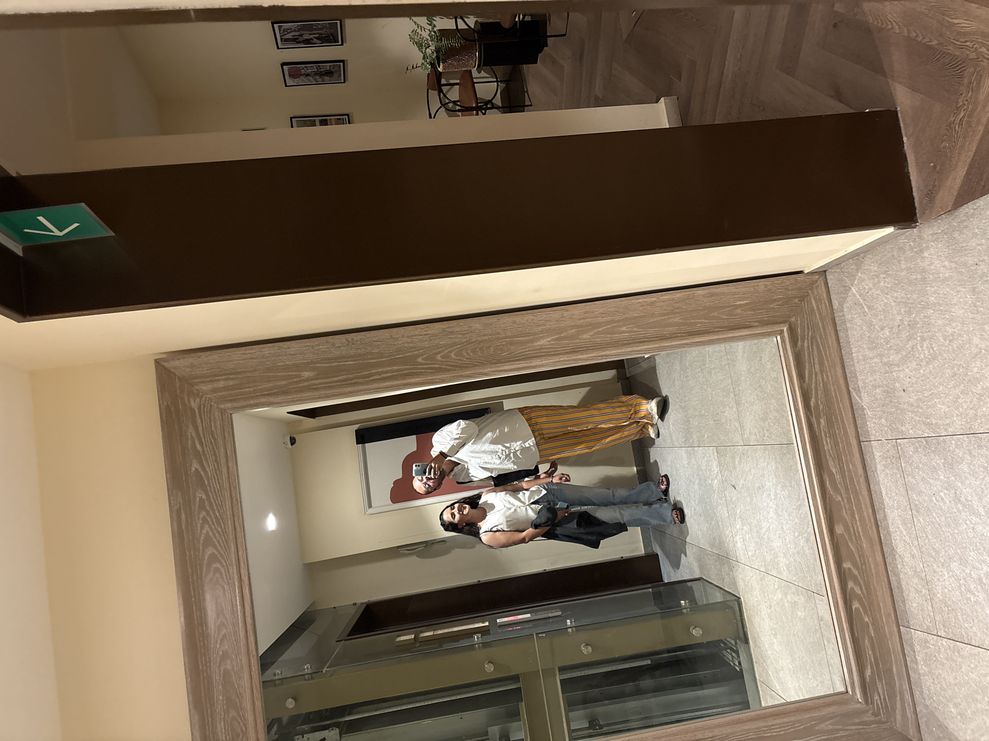 The image shows a reflection in a large mirror with a wooden frame. The reflection captures two people standing in what appears to be a building or establishment, possibly in Mexico City, Mexico.
The person in the reflection wears a white shirt, jeans, and has a black bag slung over their shoulder. A second person stands next to the first, wearing a white shirt and striped pants. The second person is holding a phone, seemingly taking the picture.
Behind the subjects is a glass elevator or a clear barrier with horizontal lines. The floor appears to be tiled. The image also reflects part of the ceiling, including a light fixture and a sign with a green arrow. The mirror reflects a hallway with art hanging on the wall and a potted plant on a table.
