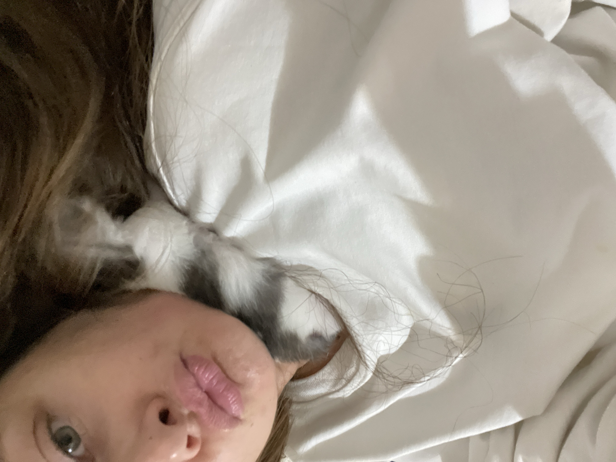 The image shows a close-up of a person's face, with their eyes and lips prominently featured. The person appears to be lying down, possibly in bed, as indicated by the soft white fabric that fills much of the frame. A portion of a small, grey and white animal, possibly a kitten, is visible nestled near the person's head and neck, peeking out from under the fabric. The lighting suggests it might be daytime, possibly indoors. The overall mood conveyed is intimate and relaxed, with a sense of comfort and closeness between the person and the animal. There is no visible text in the image.