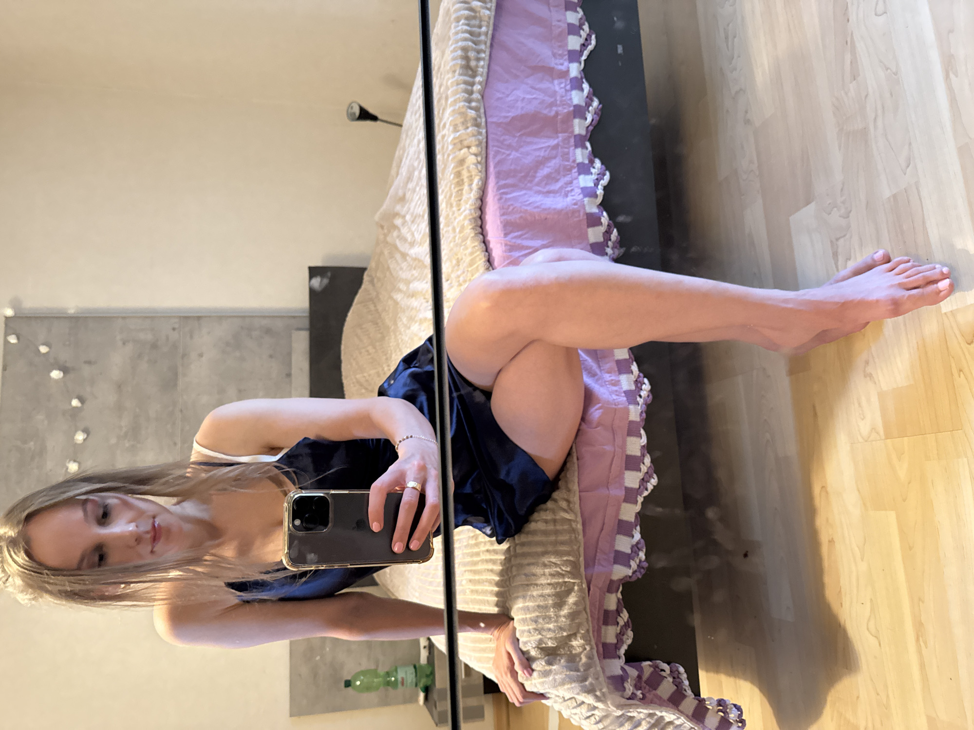 A woman is sitting on a bed, holding a phone in her right hand, and taking a mirror selfie. She is wearing a dark blue slip dress with thin straps and a white bra underneath. Her legs are crossed. The bed has a beige textured comforter and a pink bed skirt with a white and purple trim. In the background, there is a wall with a textured gray panel and some decorative lights. To the left of the bed, there is a nightstand with a green bottle on it. The floor is made of light-colored wood. The image appears to be taken indoors, in a bedroom.