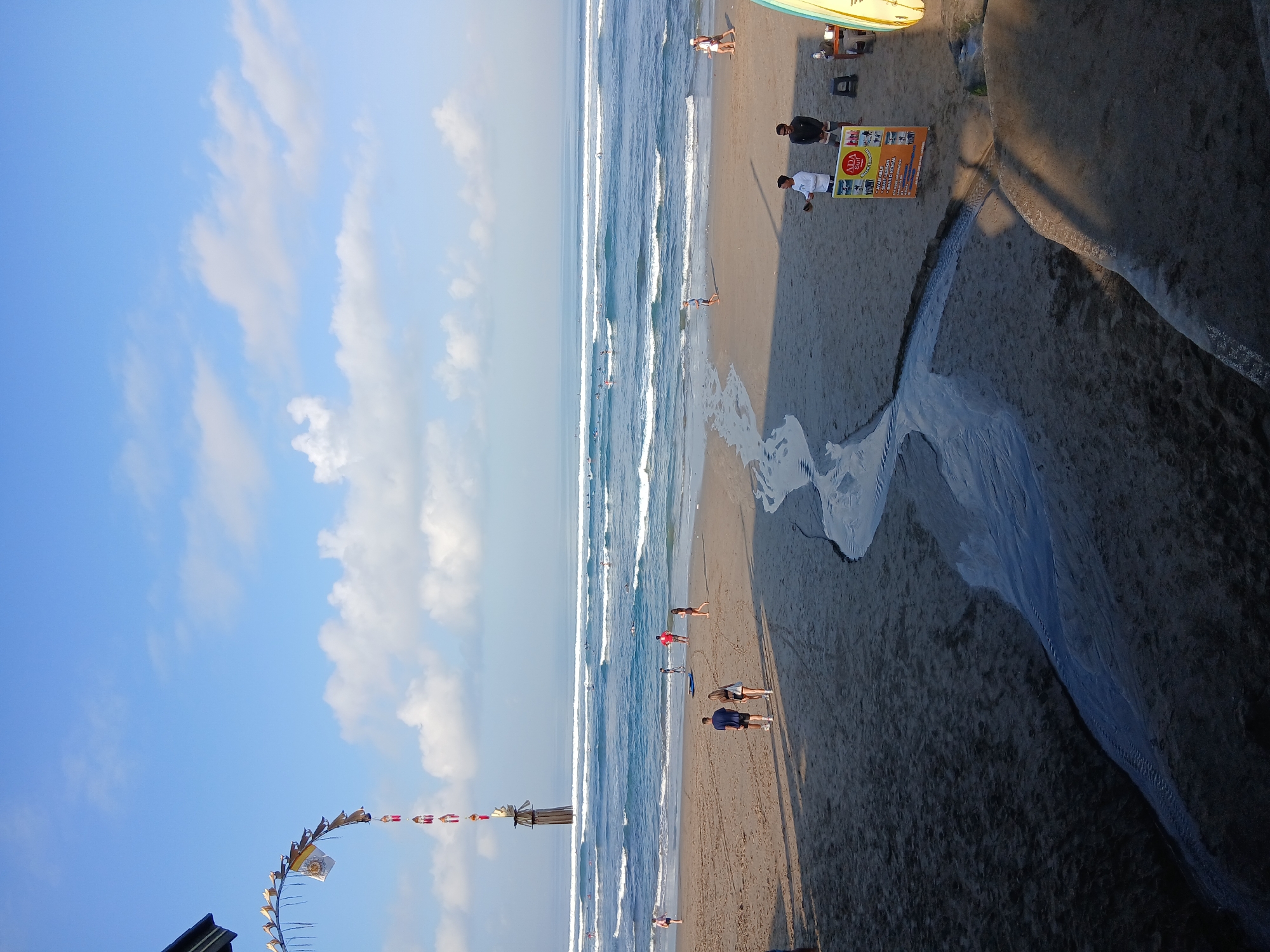 This is an eye-level, vertical, medium shot of Kuta Beach in North Kuta, Indonesia. The image captures a serene beach scene under a bright blue sky dotted with scattered white clouds. In the foreground, a small stream of water meanders across the sandy beach. Several people are scattered across the sand, some walking, others standing. In the middle ground, the ocean stretches out, with gentle waves breaking and numerous surfers visible in the water. Further back, a long, decorative banner, likely a Penjor, arcs gracefully from the left side of the frame towards the sea. Two men stand near a sign advertising surf lessons and board rentals on the right side of the image. The overall atmosphere is peaceful and leisurely, conveying a typical day at a popular beach destination.