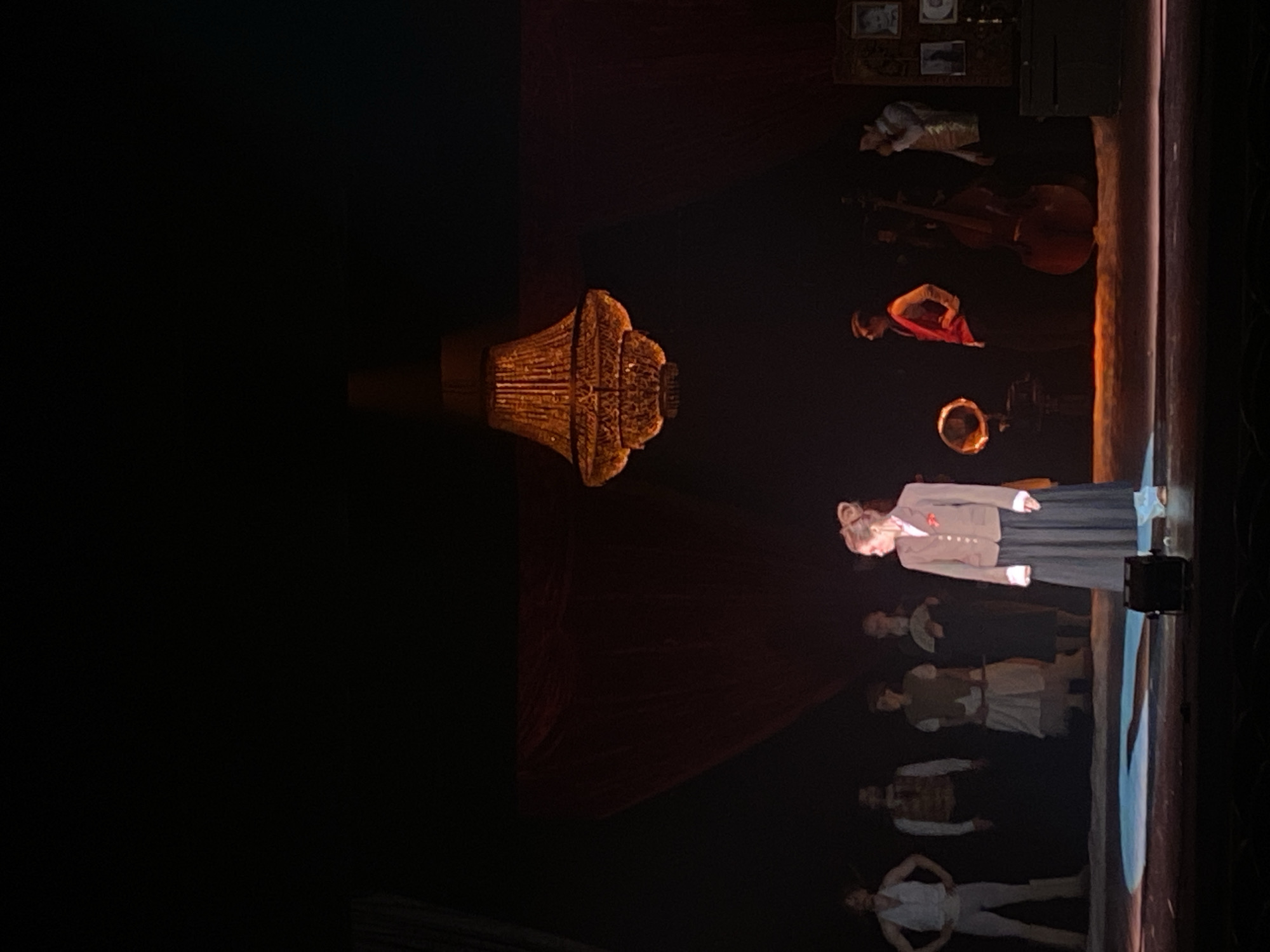 A wide shot of a theatrical production. On stage are multiple actors wearing period costumes. Above them hangs an ornate chandelier. To the side and behind the actors, musicians and their instruments are visible. The background is dark. The location is a stage in a theater in the city of Шебунино, Russia.