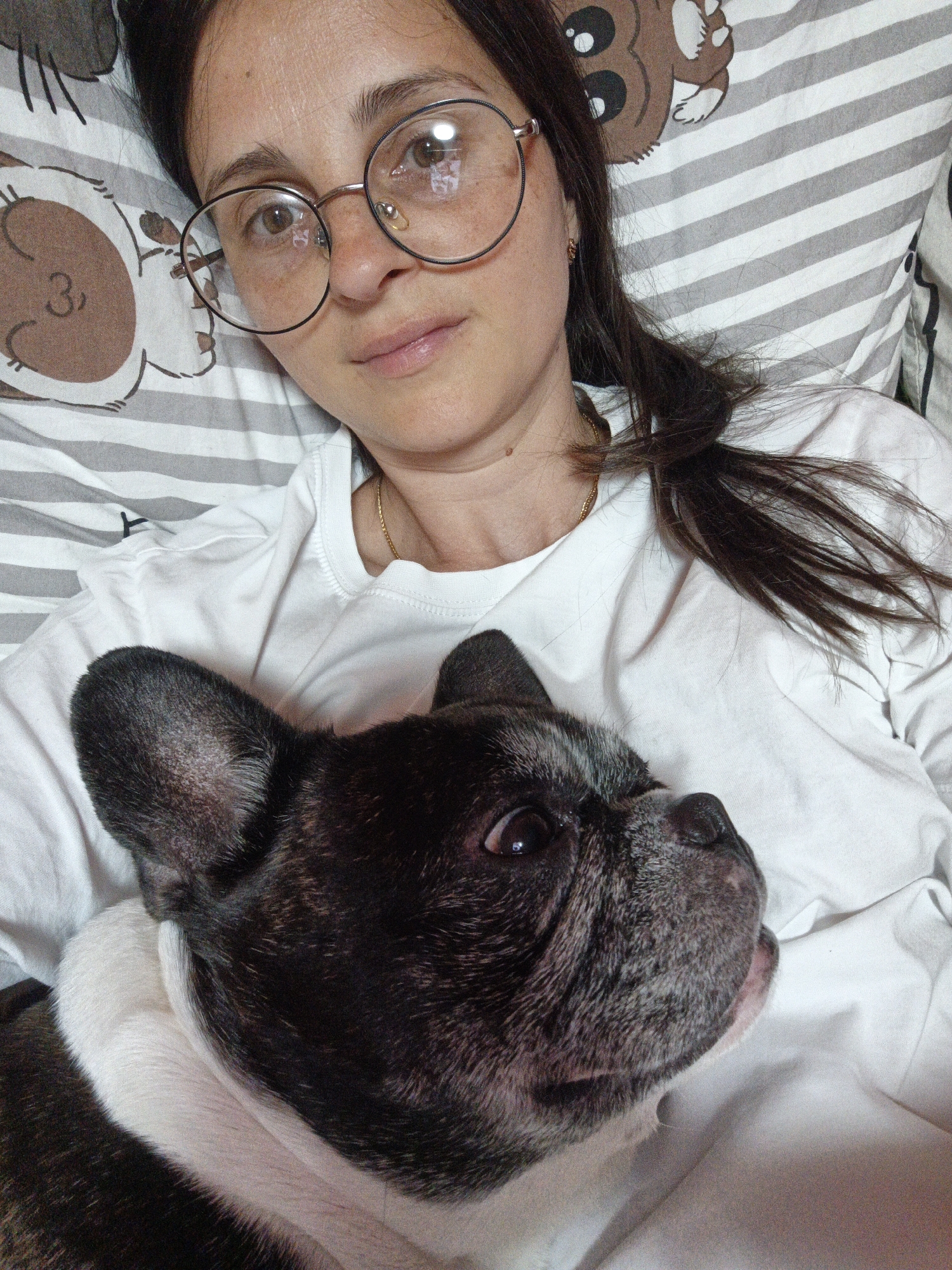 A woman wearing round glasses and a white t-shirt lies in bed with a French Bulldog. The woman has dark hair and is looking towards the camera with a neutral expression. The dog is black and white and is nestled against her chest, looking to the side with its mouth slightly open. The bedding has a pattern of gray and white stripes, with a graphic of a bear face on the pillow. The scene appears to be indoors, likely in a bedroom. The lighting suggests it could be daytime. There is no visible text in the image.