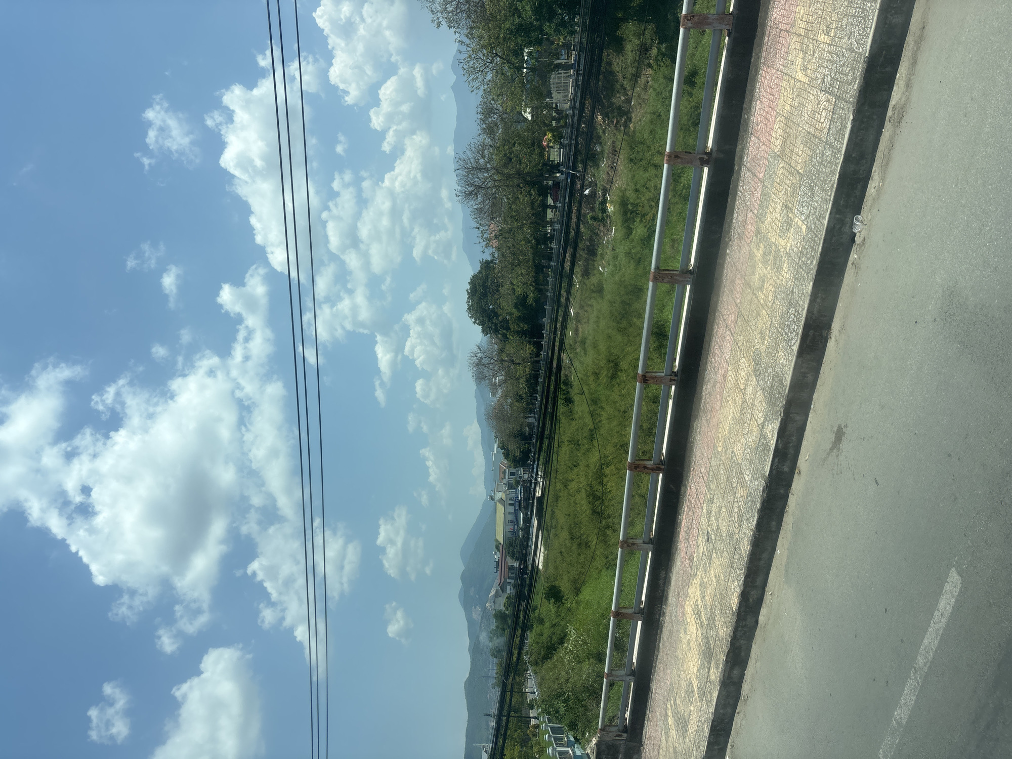 The image depicts a landscape scene under a blue sky with scattered white clouds. In the foreground, there is a paved road with a white line, bordered by a sidewalk with patterned tiles and a metal railing. Beyond the sidewalk, there is a grassy embankment leading down to a vegetated area with some debris, followed by a set of train tracks. Further in the distance, there are trees, buildings, and rolling hills or mountains. Power lines are visible crisscrossing the sky. The scene appears to be an outdoor, possibly semi-urban or suburban, environment.