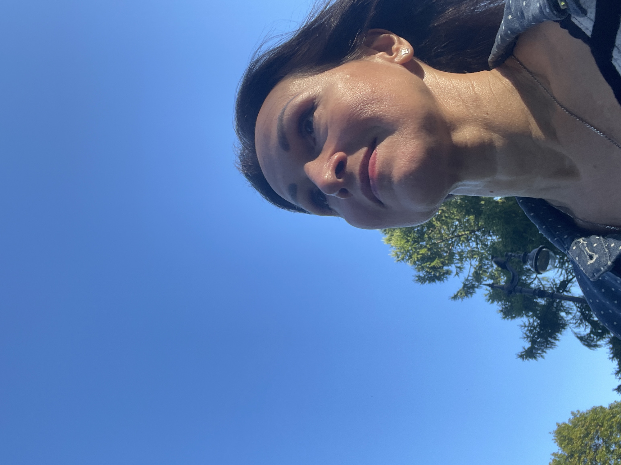 The image shows a woman from the chest up, looking upwards towards a clear blue sky. She has dark hair and is wearing a dark blue patterned shirt. A silver necklace is visible around her neck. In the background, green trees and a lamppost are visible. The lighting suggests it is daytime. The context provided suggests this image was taken in the city of Saint Petersburg, Russia.