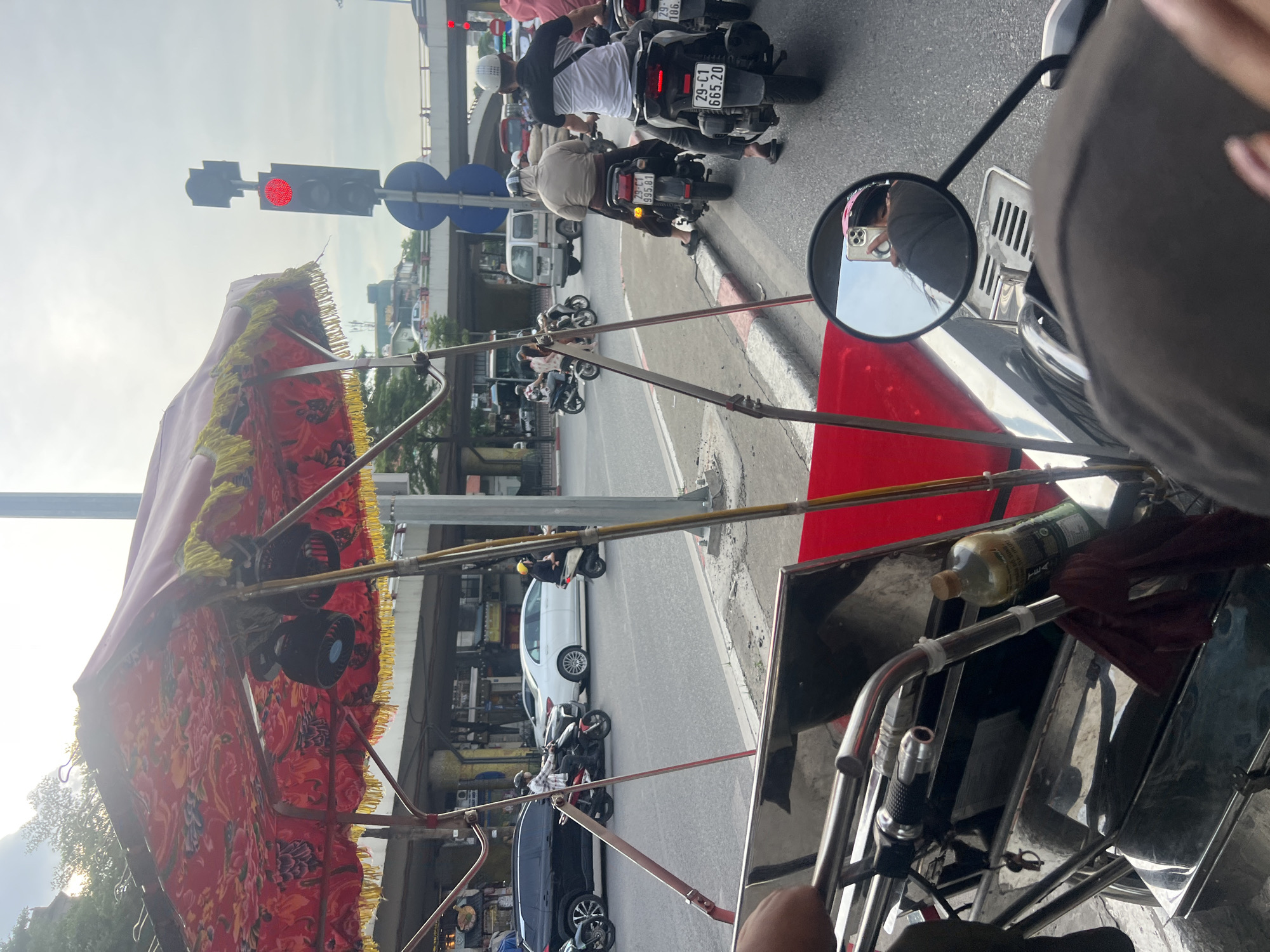The image captures a street scene in Hanoi, Vietnam, from the perspective of someone riding in a cyclo. The foreground features the handlebars and part of the cyclo's red seating area, along with a metal frame and a bottle of liquid. In the mid-ground, traffic moves along a paved road. Several motorbikes are visible, with riders wearing helmets and casual clothing. A black SUV and a white sedan are also present in the traffic. A prominent traffic light shows a red signal. The background reveals an elevated road or bridge structure, and buildings lining the street. The sky is overcast. The scene depicts a typical urban traffic environment in Hanoi, characterized by the prevalence of motorbikes and the presence of cyclos as a mode of transport.