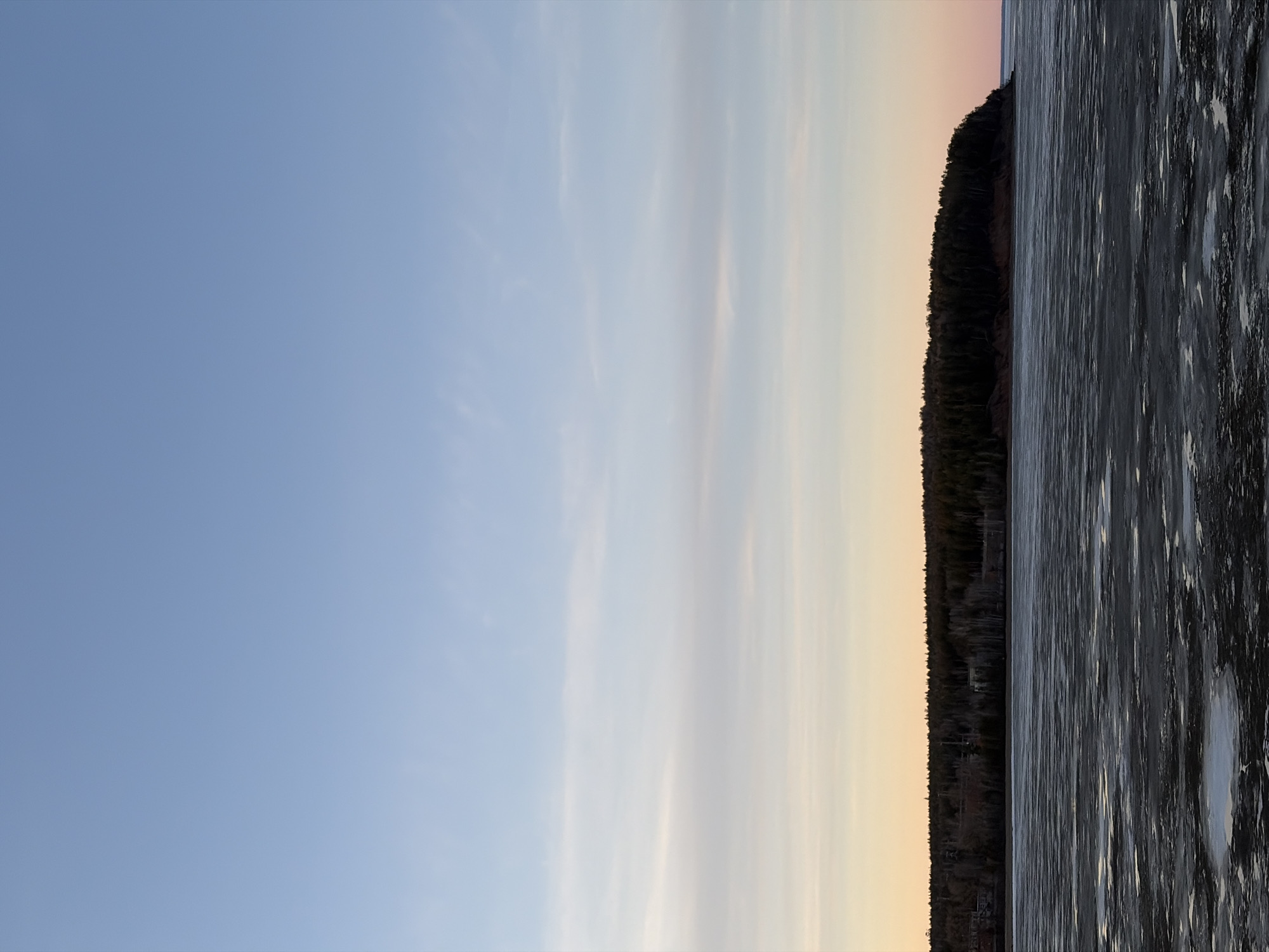 The image shows a landscape scene. The left side of the frame is dominated by a blue sky with wisps of white clouds, transitioning to a warm, pale yellow-orange near the horizon. On the right side, a dark treeline borders the edge of a large body of water with a textured surface, possibly reflecting light or containing ice formations. The presence of the body of water could suggest the location is in the city of Chaikovsky, Russia, as it is located on the shore of the Votkinsk Reservoir. No people are visible in the image.