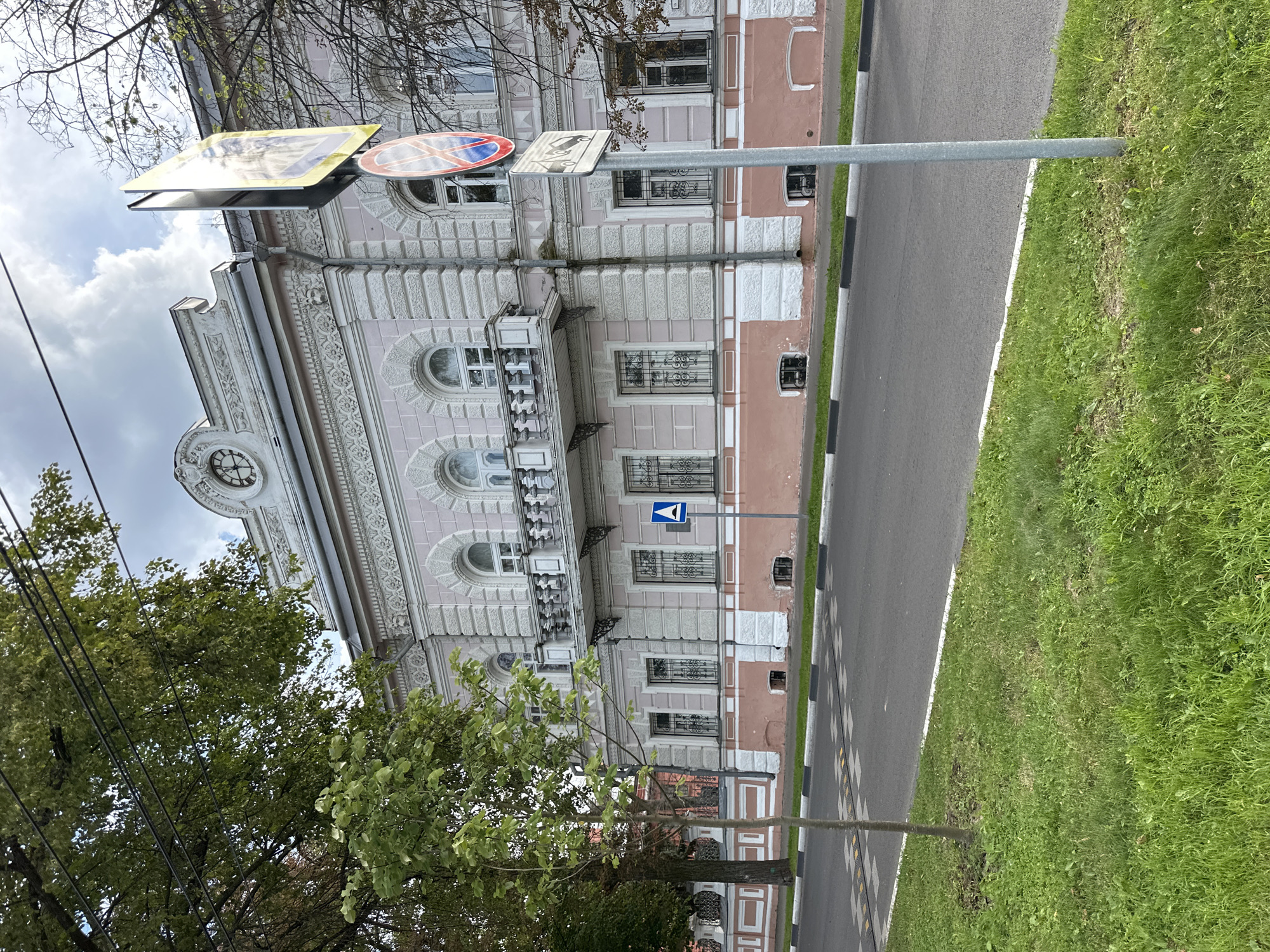 The image shows a street scene in the city of Zhukovo, Russia. In the foreground, a paved road with a white line marking the edge and speed bumps is visible. To the left of the road is a strip of green grass with a young tree.
Dominating the background is a large, ornate pink and white building with classical architectural features. The building has multiple windows, some with decorative metal grates, and a balcony on the second floor supported by ornate brackets. The upper facade features decorative molding and a circular clock face.
A tall metal pole with traffic signs stands on the right side of the image, in front of the building. The signs appear to be a pedestrian crossing sign and another circular sign with red and blue markings.
The sky is partly cloudy with some patches of blue visible. The overall impression is a calm, urban setting with a focus on the historical architecture of the building. There are no people or animals visible in the frame.