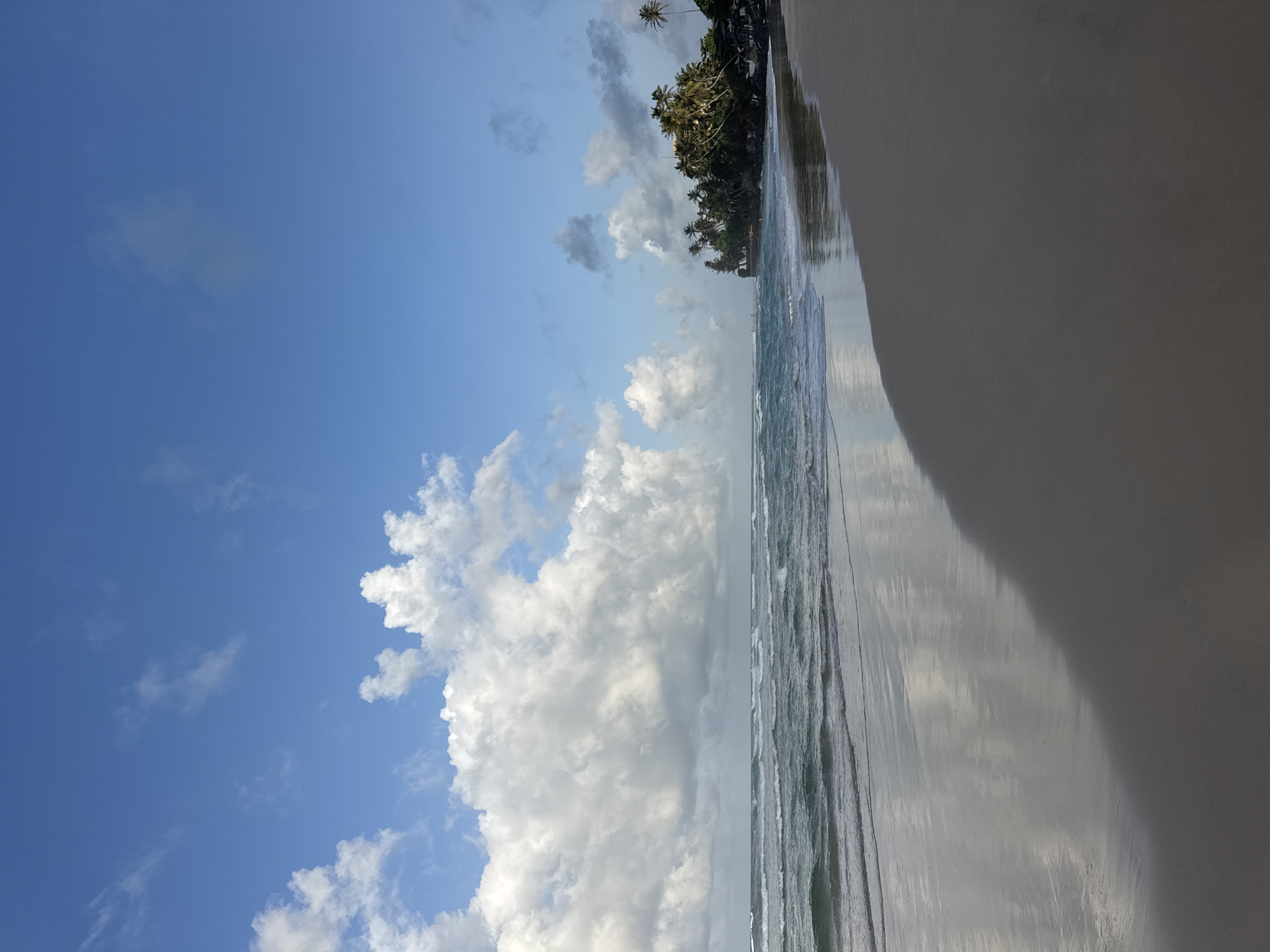 A wide shot captures a serene beach scene in Kathaluwa, Sri Lanka. The sky is a vibrant blue, dotted with fluffy white clouds on the left and lighter, scattered clouds on the right. Gentle waves roll onto a sandy shore, reflecting the sky and clouds. To the right, a lush green coastline is dotted with palm trees, with a few buildings visible nestled amongst the foliage. The sand in the foreground is wet and reflects the sky, creating a mirror-like effect. No people are visible in the frame.