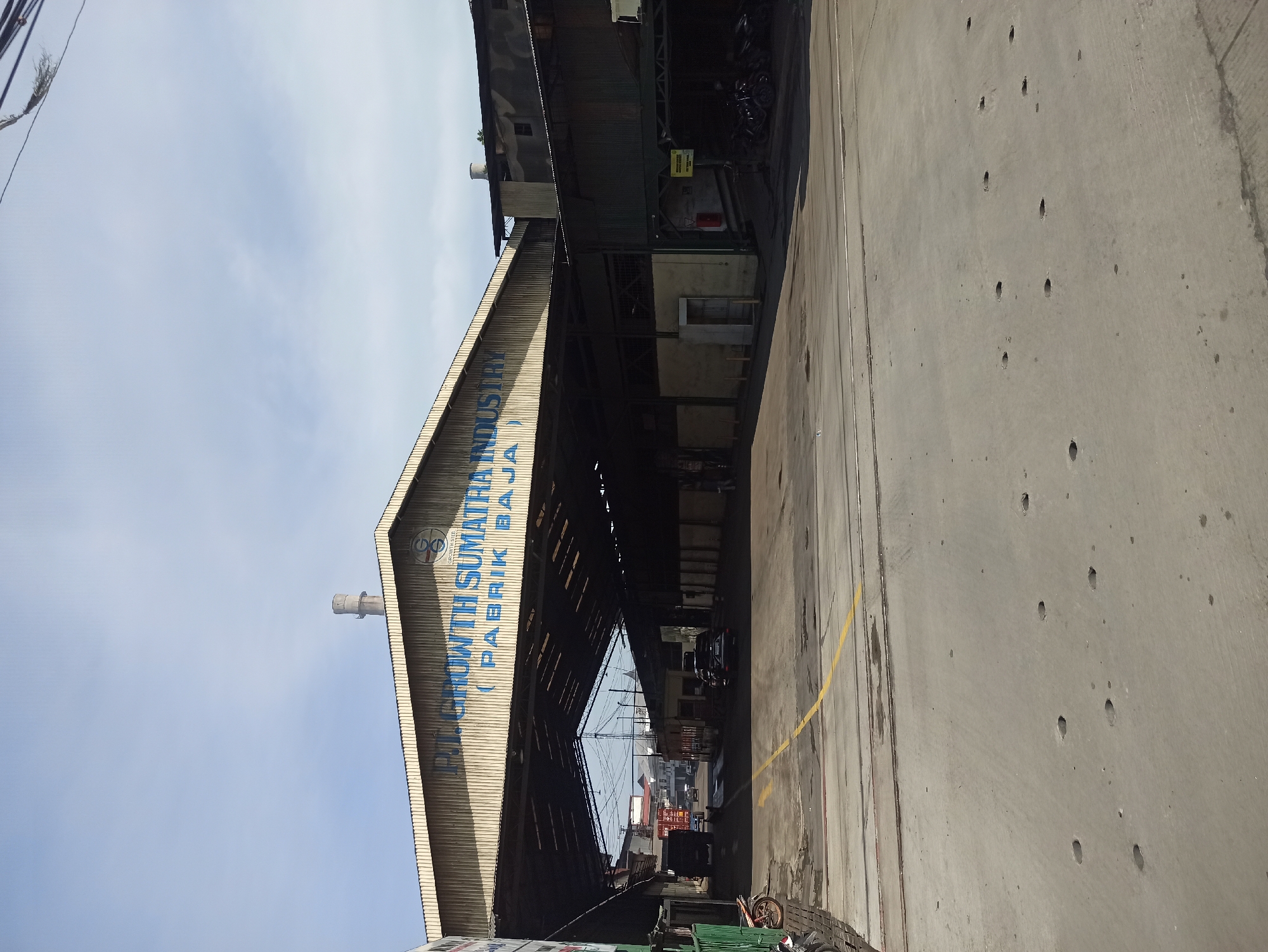 The image shows the entrance to PT. Growth Sumatra Industry (Pabrik Baja) in the city of Medan, Indonesia. The factory's name is prominently displayed in blue lettering on the arched structure above the entrance. The scene depicts an industrial area with a large, open-sided warehouse or factory building. The ground is paved with concrete and shows some wear and tear, including potholes. Yellow lines are painted on the ground, possibly indicating traffic lanes or safety zones.
In the background, several vehicles are visible, including trucks and cars, suggesting ongoing industrial activity. A small booth or security post is situated near the entrance. The sky is partly cloudy. To the left of the entrance, a motorcycle is parked. The overall impression is one of a working industrial facility.