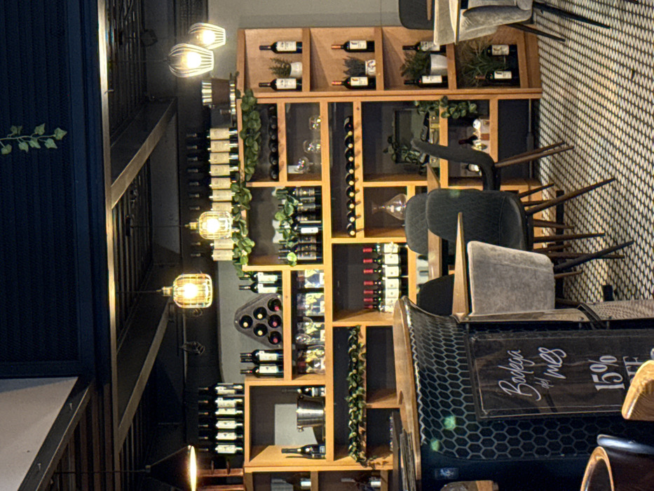 A restaurant interior showcases shelves packed with wine bottles, accented by decorative greenery. The shelves are part of a wooden structure, possibly a wine display. In front of the shelves, several chairs with wooden legs are stacked, one featuring a visible 'Bodega del Mes 15%' sign on its back. The room is illuminated by industrial-style pendant lights.