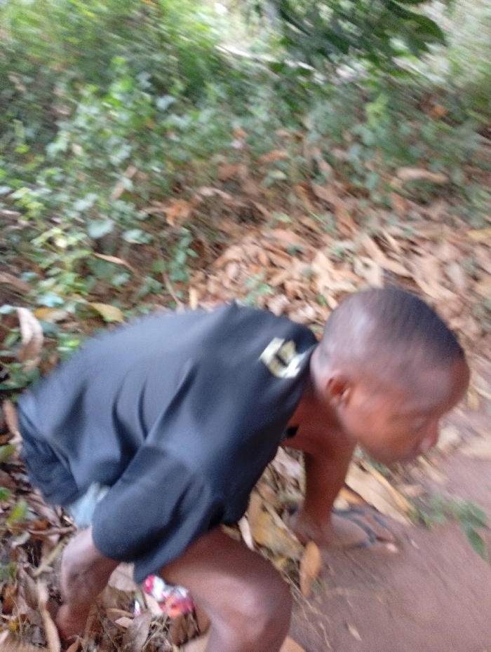 The image depicts an individual, identified as a male, in a crouching or bending posture outdoors in Eziudo, Nigeria. The subject wears a dark, long-sleeved shirt featuring a light-colored emblem on the upper right sleeve. Below the shirt, a glimpse of patterned undergarment or shorts is visible. His head, with short dark hair, is turned to the right and appears blurred due to motion. A pair of dark flip-flops rests on the reddish-brown soil near his right foot. The ground is largely covered with dry, fallen leaves and some sparse green vegetation. The background consists of heavily blurred green foliage, indicative of a natural, vegetated environment. The overall image exhibits motion blur, suggesting movement by the subject or the camera at the time of capture.