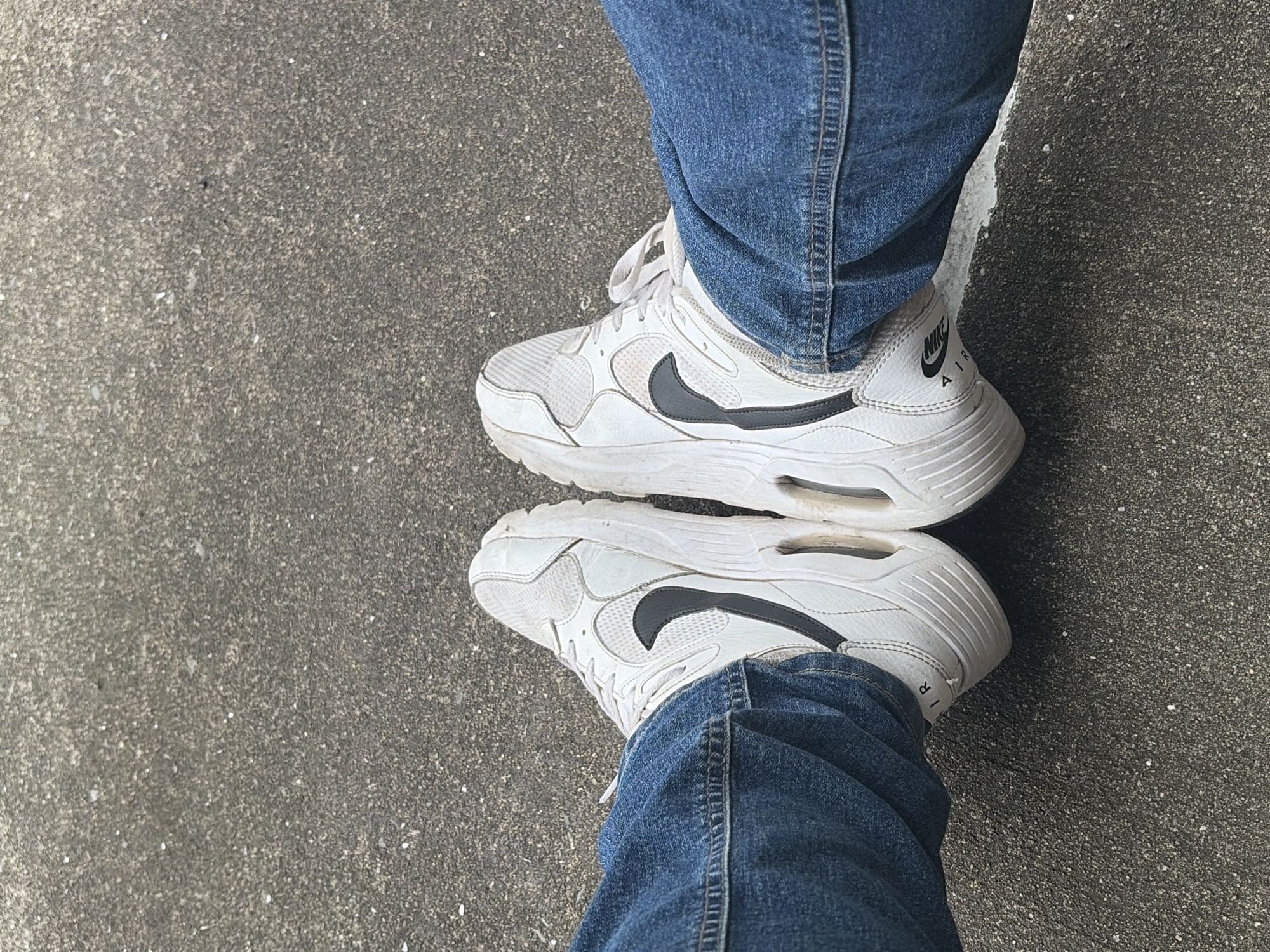 The image shows a view of a pair of white Nike Air sneakers and denim jeans on an asphalt surface. The shoes are slightly dirty. The location is indicated as Edison, United States of America, but there are no specific details in the image to confirm this.