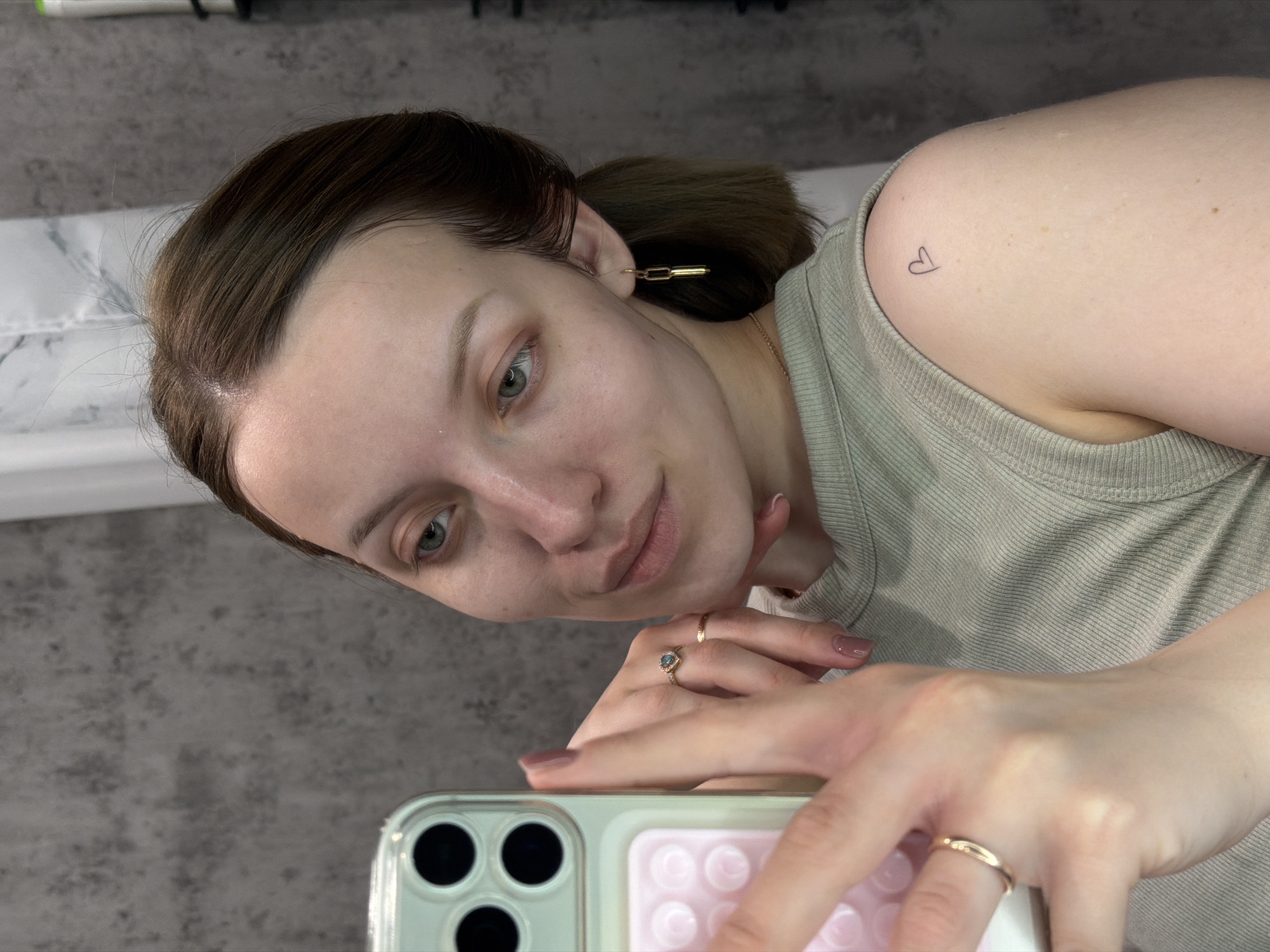 A light-skinned woman is holding a smartphone in front of her face, seemingly taking a selfie. The phone case is light green with a pink, bubble-like pattern. She is wearing rings on her fingers; one is a gold band, and the other appears to have a light-colored stone in the center. Her hair is dark brown, pulled back, with a gold chain earring. She is wearing a light-colored tank top, and a small heart tattoo is visible on her shoulder. The background includes a grey wall and a white surface. There is no indication of the location being Reshetnikovo, Russia.