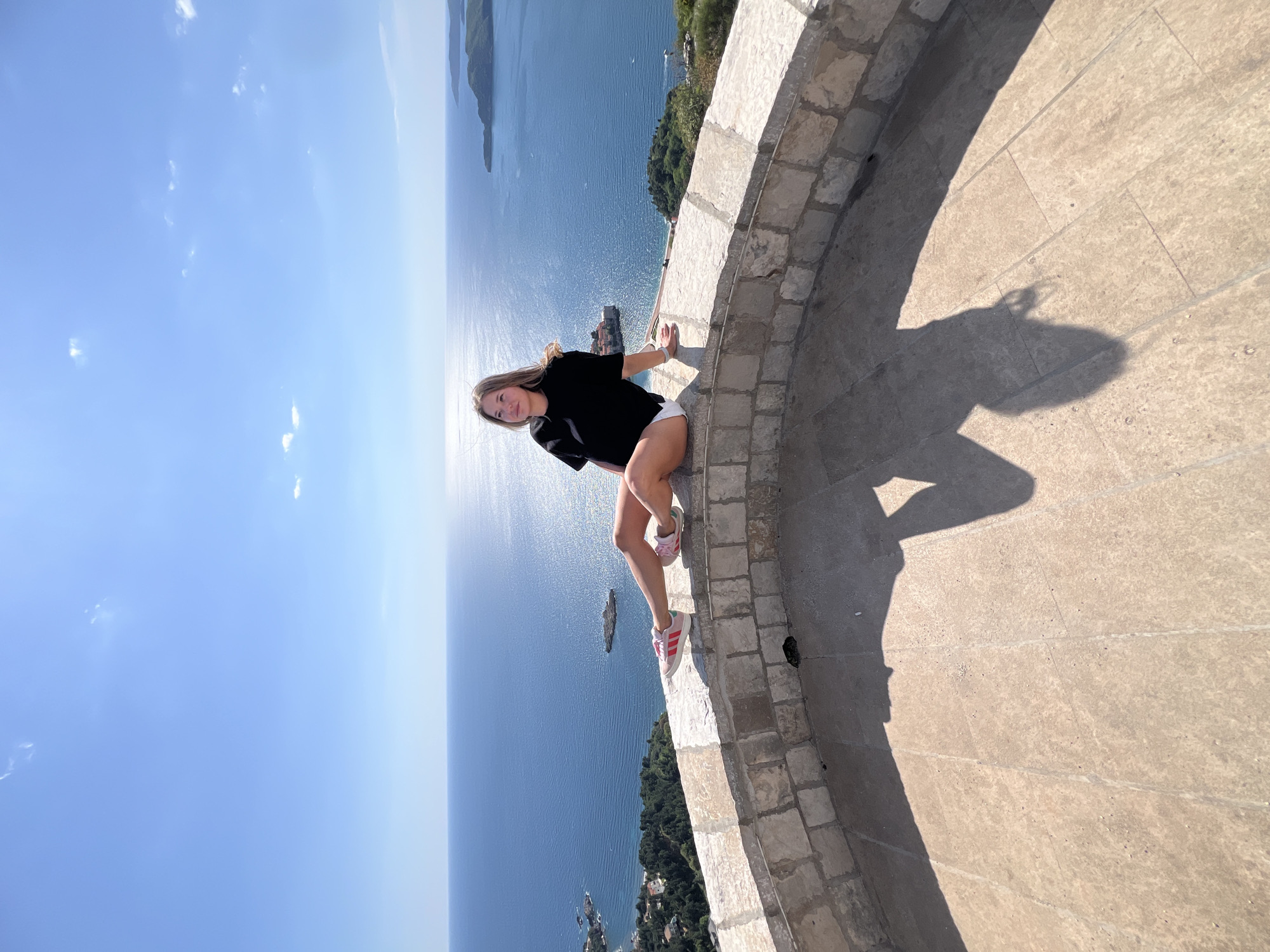 A young woman is captured posing on a curved stone wall, overlooking a vast expanse of the Adriatic Sea. The image is oriented vertically, rotated 90 degrees counter-clockwise from its likely original portrait capture. The woman, with long, light-brown hair, wears a black t-shirt, white shorts, and pink and white sneakers. She sits with bent legs, her hands resting on the stone wall, and looks directly at the camera with a subtle smile, conveying a relaxed and pleasant demeanor.
The scene is a scenic overlook, typical of coastal viewpoints in Sveti Stefan, Montenegro. Below the stone barrier, dense green foliage covers the sloped terrain. The deep blue sea extends far into the horizon, dotted with distant islands and land formations. The sky above is a clear, bright blue, accented by a few scattered white clouds. The photograph was taken in strong daylight, with the sun's glare visible in the upper left, casting prominent shadows of the wall and the woman onto the light-colored, paved ground to the right. No text is visible in the image.