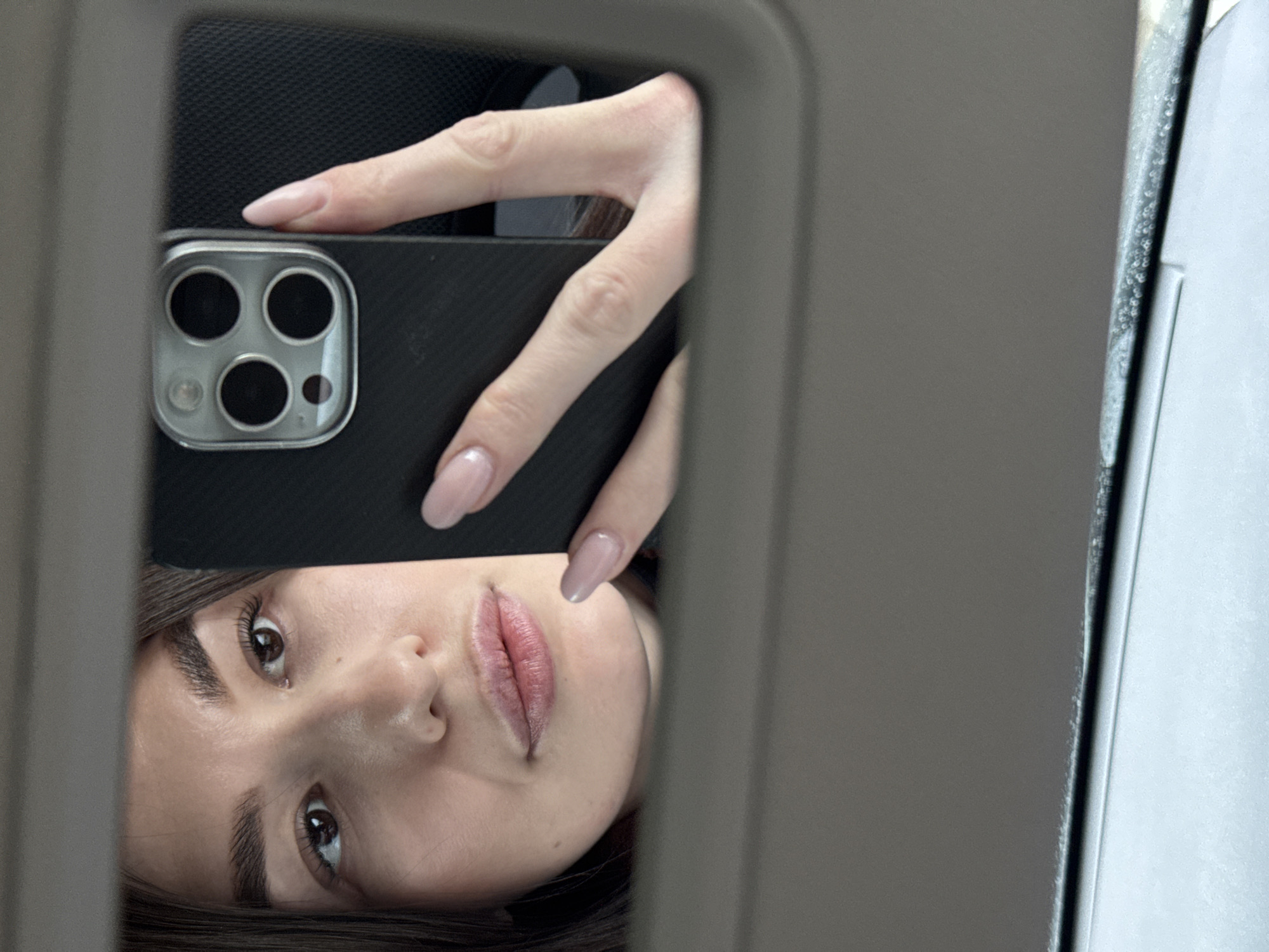 A selfie is reflected in a car's sun visor mirror. A fair-skinned woman with dark hair and brown eyes holds a black smartphone with a multi-lens camera facing inward towards her face. She has manicured nails with a light pink polish. The environment is the interior of a car, evident from the visor and the adjacent car window. No evidence exists of the city of Chaikovsky, Russia.