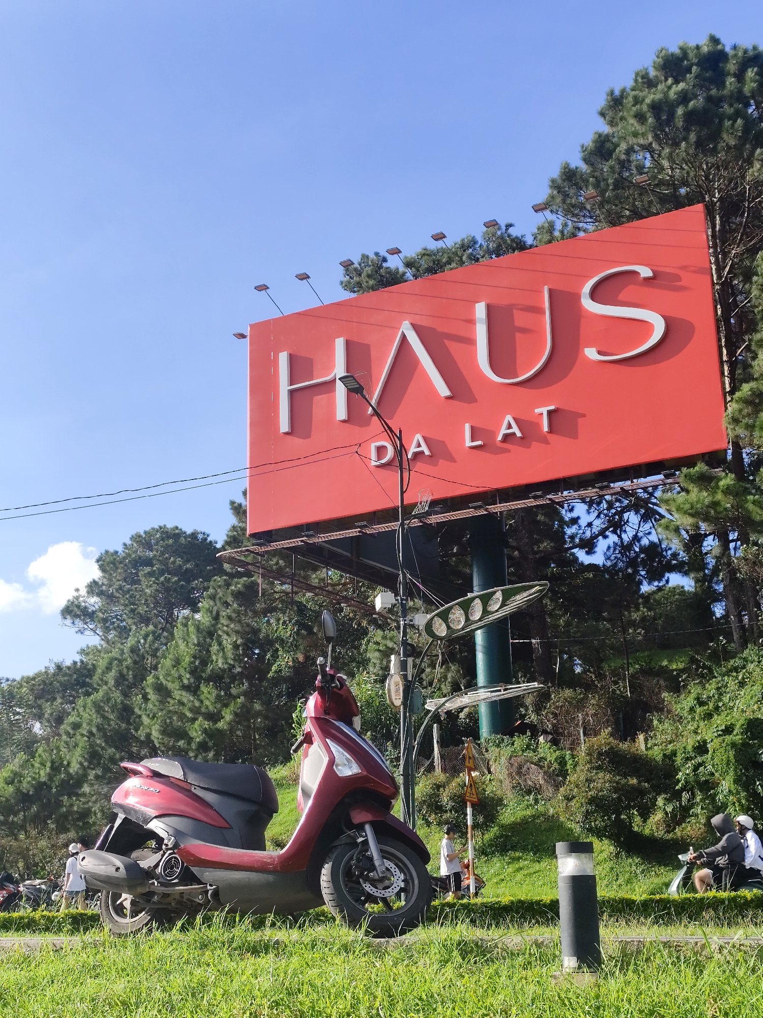 A low-angle shot captures a red scooter parked on a grassy slope in Da Lat, Vietnam. Behind the scooter, a large red billboard with white lettering reads "HAUS DA LAT." The billboard is mounted on a metal structure and is partially obscured by trees. A lamppost stands in front of the billboard. In the background, several people are visible, some on motorcycles. The sky is clear and blue, with a few scattered clouds. The overall scene depicts a roadside area in a city known for its pine trees and cooler climate.