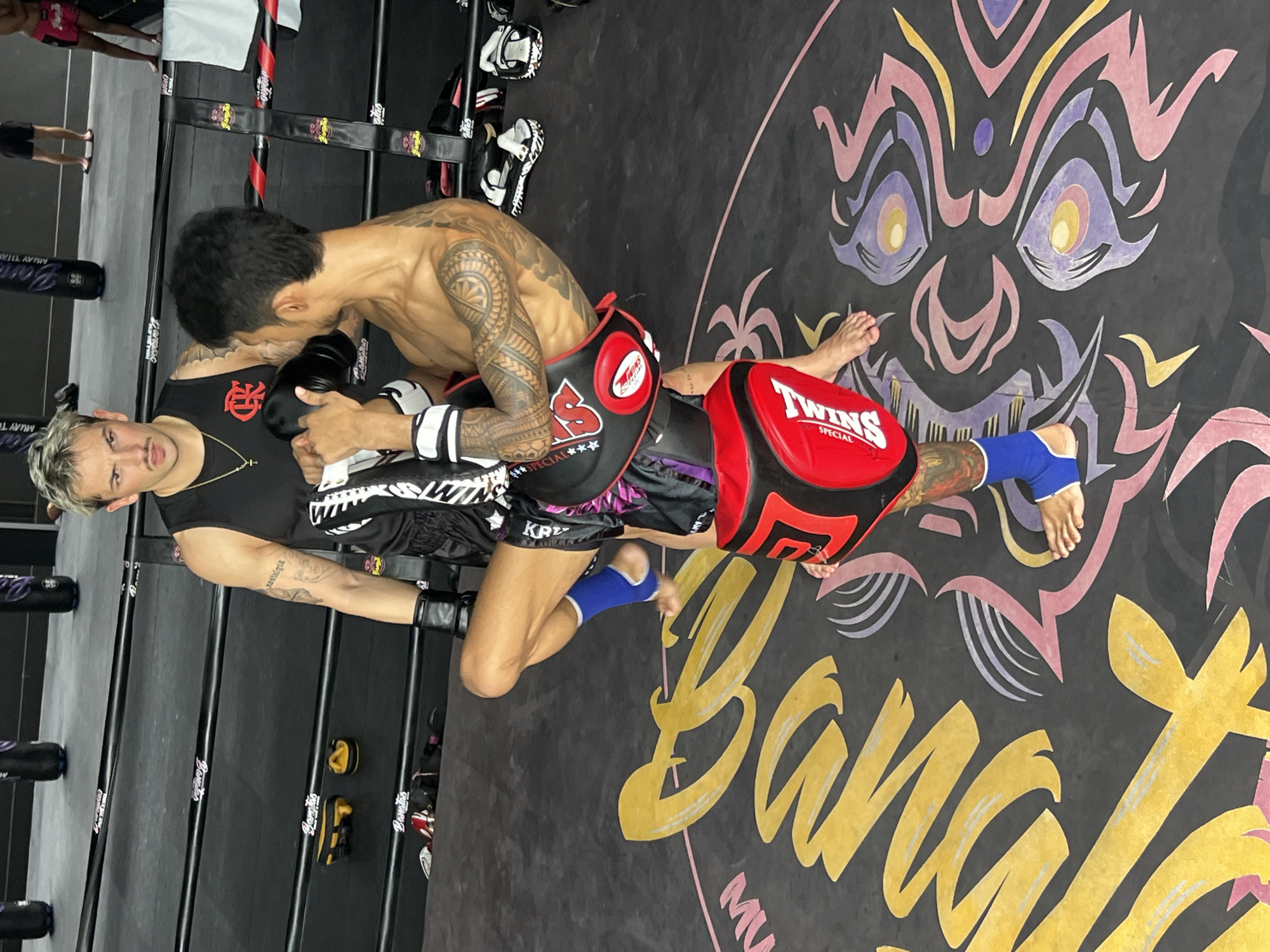 Two men are in a Muay Thai boxing ring. The man on the left has light-colored hair and a mustache, wearing a black tank top and black boxing shorts with a gold chain. The man on the right is shirtless, showing off a tattooed arm and torso, wearing black boxing shorts and holding kickboxing pads. He is actively training the man on the left. The gym floor has a large graphic of a stylized face and lettering. Visible text includes "TWINS SPECIAL," "MUAY THAI," and "BANGKOK." The scene appears to be indoors, likely during the day, given the lighting. The overall atmosphere suggests a training session in a martial arts gym. The location context is Choeng Thale, Thailand.