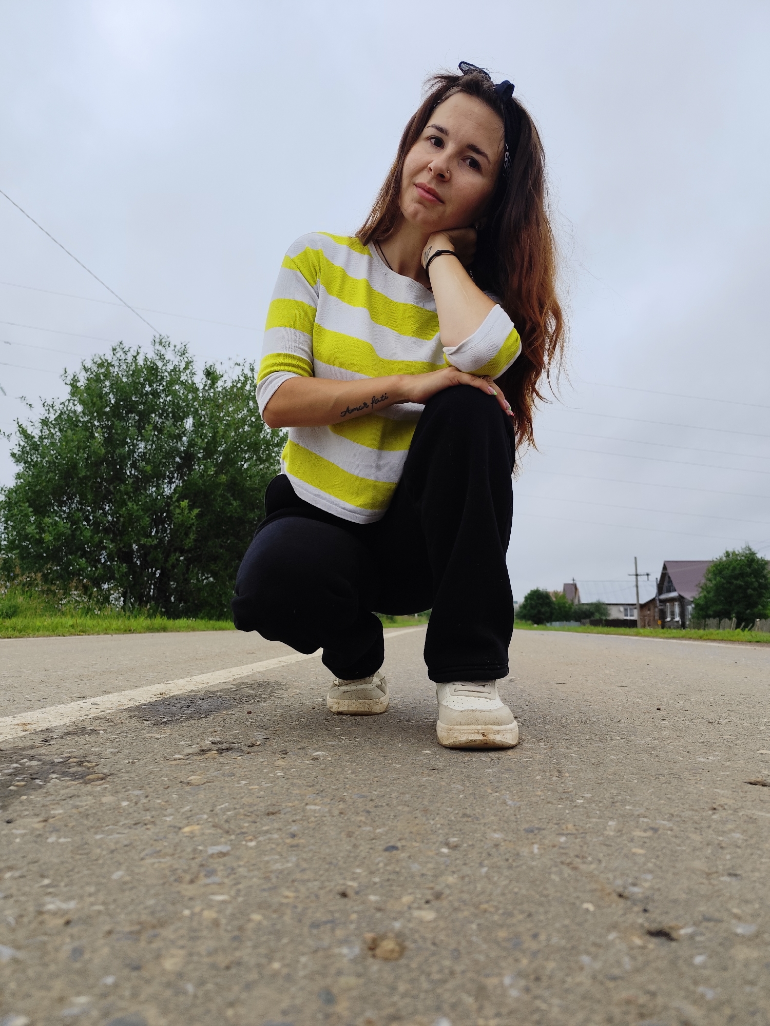 A young woman with long brown hair and a blue bandana is crouched on an asphalt road. She is wearing a yellow and white striped t-shirt and black pants, with white sneakers. Her left arm is resting on her knee, and her hand is supporting her chin. She is looking towards the camera. The road has a white line down the center. In the background, there are trees, power lines, and some houses. The sky is overcast. The location is identified as Fokino, Russia.