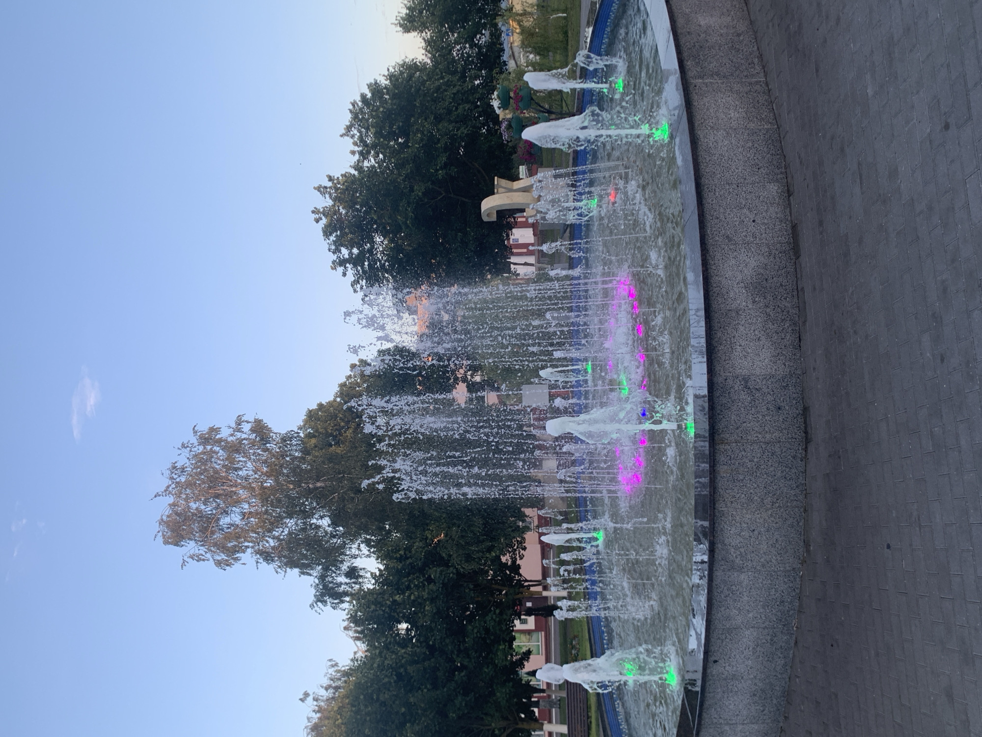 A circular fountain with multiple water jets is illuminated by green and pink lights. The fountain is surrounded by a stone base and is situated on a paved area. In the background, there are trees, buildings, and a sculpture that appears to be made of large, block-like letters. The sky is clear and blue, suggesting it is daytime. This scene is located in the city of Kapyĺ, Belarus.