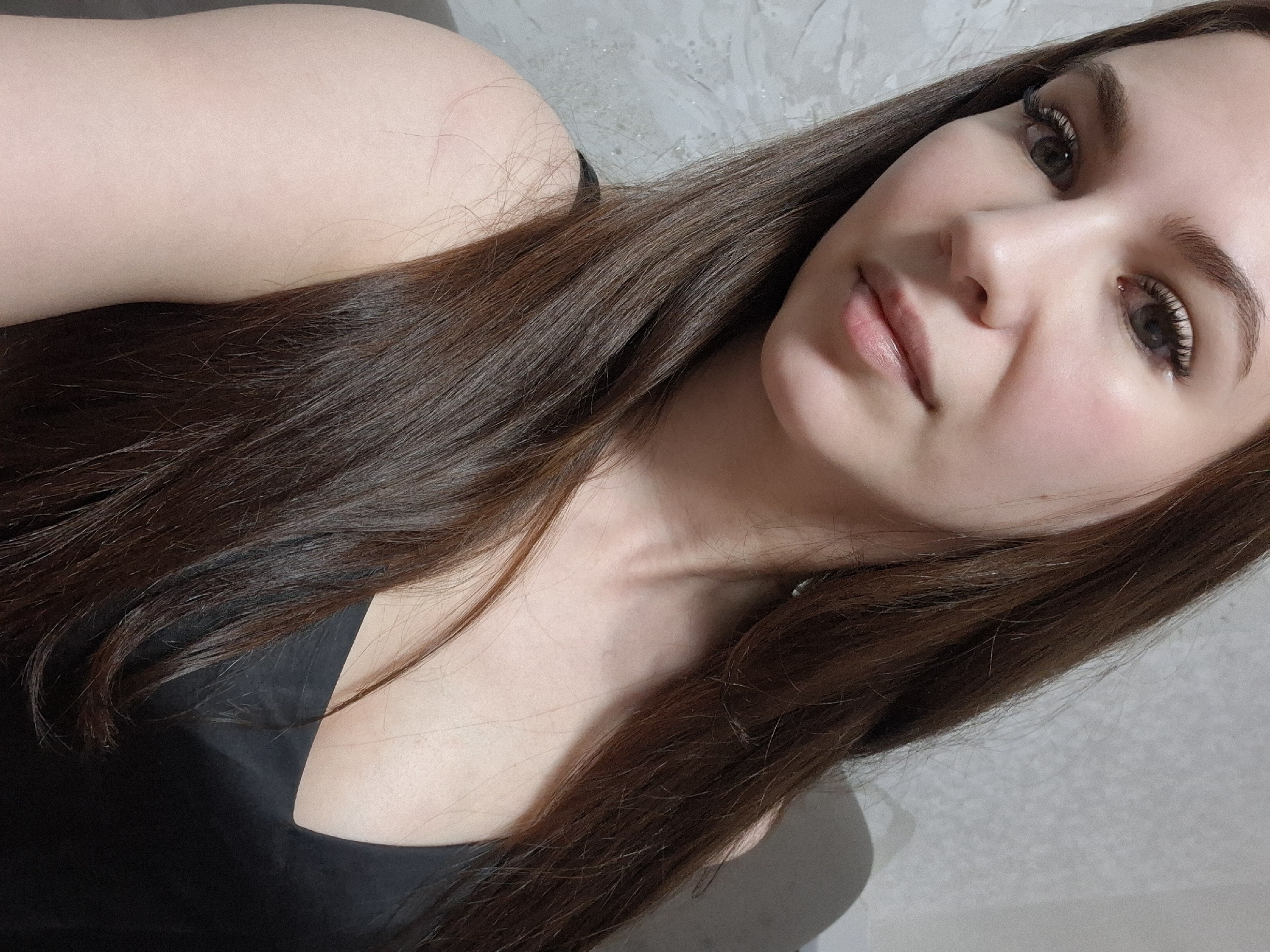 A close-up, angled selfie of a woman with long, brown hair. She has fair skin, light eyes, and defined eyebrows and eyelashes. She is wearing a black top. The background is a textured, light gray wall. The location is not discernible from the image; there is no evidence to indicate the city of Chaikovsky, Russia.