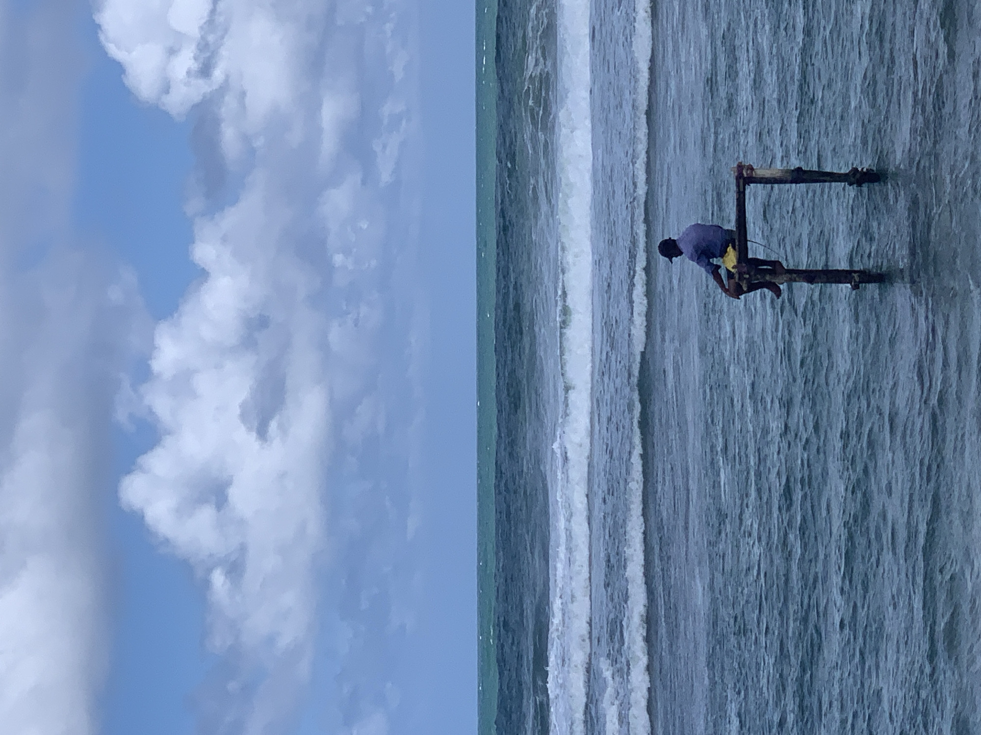 A lone man sits on a wooden structure submerged in the ocean. He is wearing a blue shirt, yellow shorts, and a dark baseball cap. The wooden structure consists of two vertical posts supporting a horizontal beam on which the man is seated. The ocean is choppy with waves breaking in the mid-ground. The horizon line is clearly visible with the sea meeting the sky. The sky is partially cloudy with a mix of blue sky and white cumulus clouds. The overall scene is one of solitude and contemplation by the sea. The location is identified as Kathaluwa, Sri Lanka.