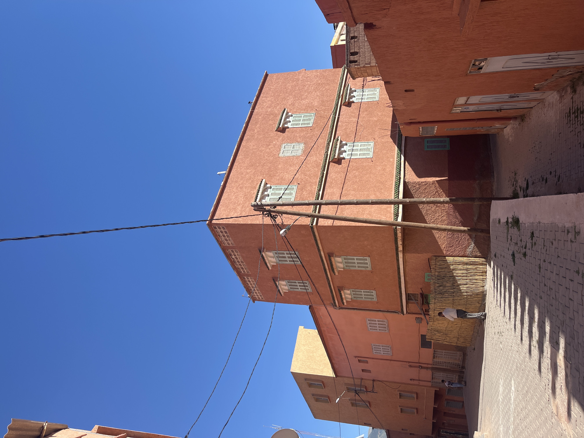 The image shows a street scene in Ouaouizert, Morocco. Tall, salmon-colored buildings line a narrow paved street. The buildings feature rectangular windows with white shutters. Utility poles and power lines are visible. A person walks on the right side of the street, casting a shadow. The sky is clear and blue. A woven bamboo-like material is attached to the side of one building.
