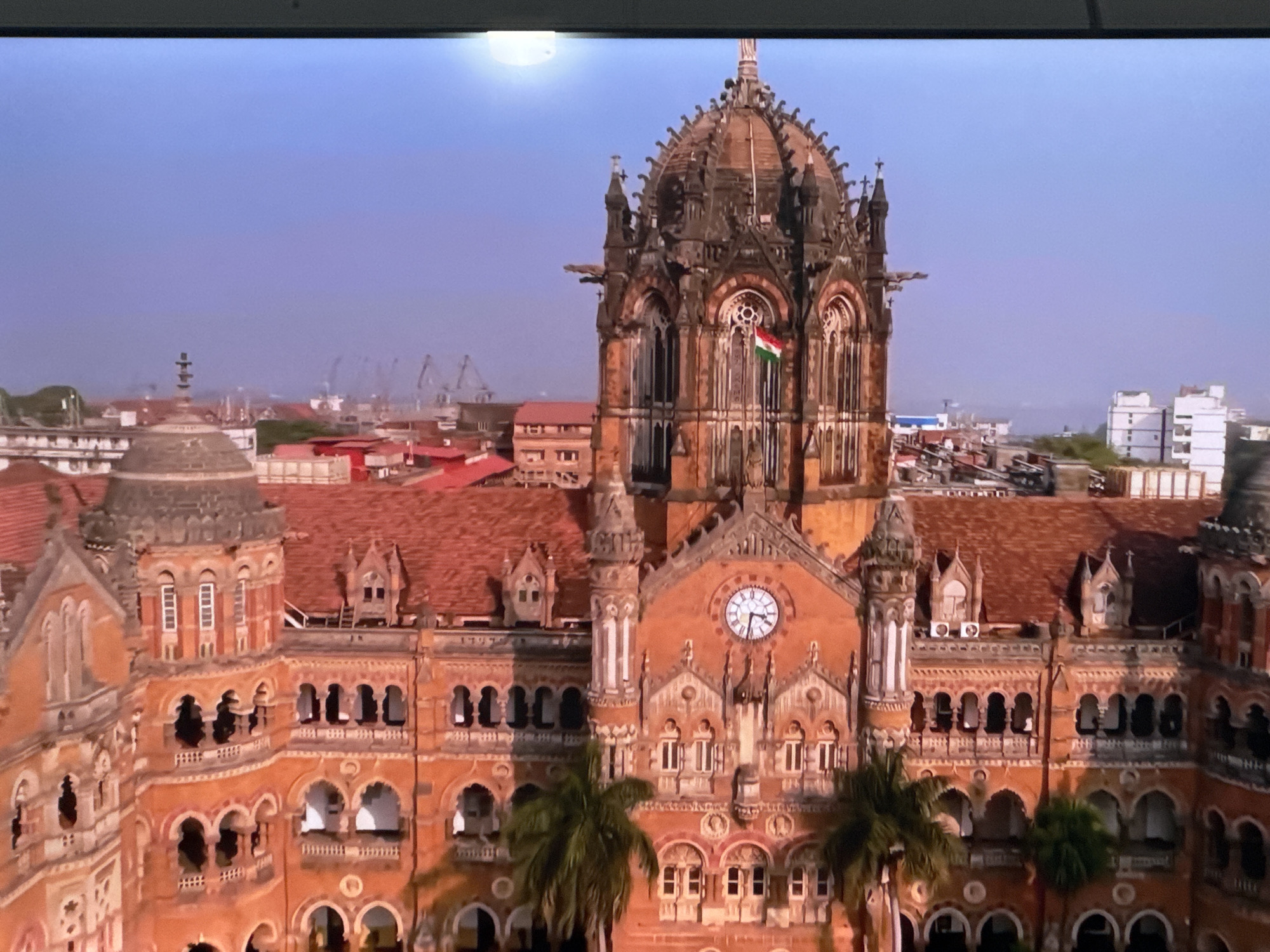 The image is an aerial view of the Chhatrapati Shivaji Maharaj Terminus (CSMT) in Mumbai, India. The magnificent Victorian Gothic structure, built with red sandstone and marble, dominates the foreground. The building features intricate architectural details, including arches, turrets, spires, and a prominent clock tower. The Indian flag is visible flying from the building's upper section. Palm trees are scattered in front of the building. In the background, a cityscape with various buildings, cranes, and a body of water can be seen. The sky is clear with a soft gradient from light blue to pale purple. There are no people or animals clearly visible in the image. The location is definitively Mumbai, India, and not Ləcət, Azerbaijan.