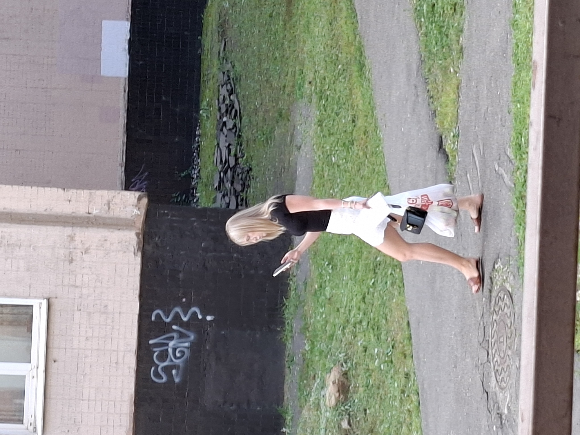 An outdoor, eye-level medium shot captures a blonde woman walking on a paved path in front of a building with a dark brick base and a lighter colored upper facade. The woman is wearing a black short-sleeved shirt and a white skirt. She is holding a smartphone in her right hand, looking down at it as she walks. In her left hand, she carries a white plastic shopping bag. A small black purse hangs from her left hand, nestled within the shopping bag. Her left leg is extended forward, and her right leg is behind her. She is wearing brown flat sandals. The path is cracked and worn, with a metal manhole cover visible on the ground. To the left of the path, there is a patch of green grass with scattered rocks. Behind the woman, the building has graffiti in white paint on the dark brick base. The background shows more grass and some dark, flat stones arranged on the ground. The lighting suggests it is daytime.