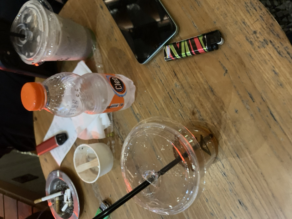 The image shows a wooden table with a variety of objects on it, including a plastic cup with a black straw, a plastic cup containing what appears to be iced coffee, a bottle of water, a small plastic cup, a lighter, a black cell phone, a red and black vaporizer, a pack of cigarettes in a plastic ashtray, and a white napkin. The table is made of wood with a distinct grain pattern. The location is Malang, Indonesia, based on the provided coordinates. The table is situated in an outdoor setting with a slight blur of grass or other greenery visible in the background. The image is taken from a bird's-eye perspective, looking down on the table and its contents. The table appears to be a typical outdoor table for a cafe or restaurant. The objects on the table suggest that people have been sitting at the table recently. The cell phone, lighter, and vaporizer suggest that the people were perhaps enjoying a break or a social gathering. The image depicts a typical scene of a cafe or restaurant setting in Malang, Indonesia.