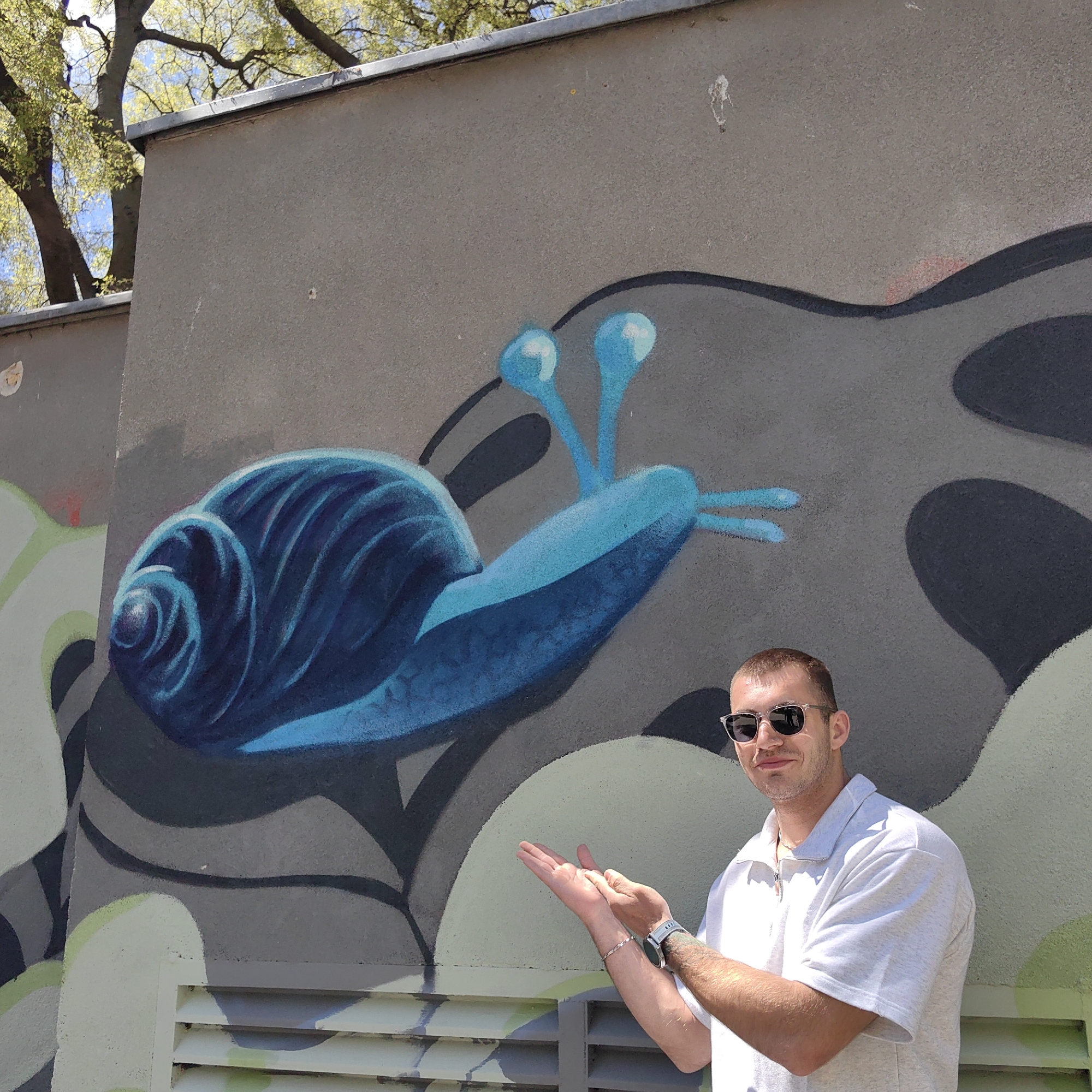 A man stands smiling in front of a graffiti-adorned concrete wall on a bright, sunny day in Varna, Bulgaria. He has short brown hair, is wearing clear-framed sunglasses, a light grey or white short-sleeved polo shirt, a silver-toned watch on his left wrist, and a thin silver bracelet on his right. He gestures with both hands, palms open and slightly upward, appearing to present or highlight the large blue snail graffiti behind him.
The graffiti depicts a stylized snail predominantly in shades of blue, with its spiraling shell and body featuring lighter and darker tones to create depth. Its eye stalks are also a bright blue. The snail is painted on a grey wall, which is further decorated with abstract, flowing patterns in various shades of grey, light green, and white, creating a dynamic backdrop. Above the wall, the green foliage of trees and a clear blue sky are visible, suggesting an outdoor urban or park setting. Part of a ventilation grate can be seen at the bottom of the wall. There is no visible text in the image.