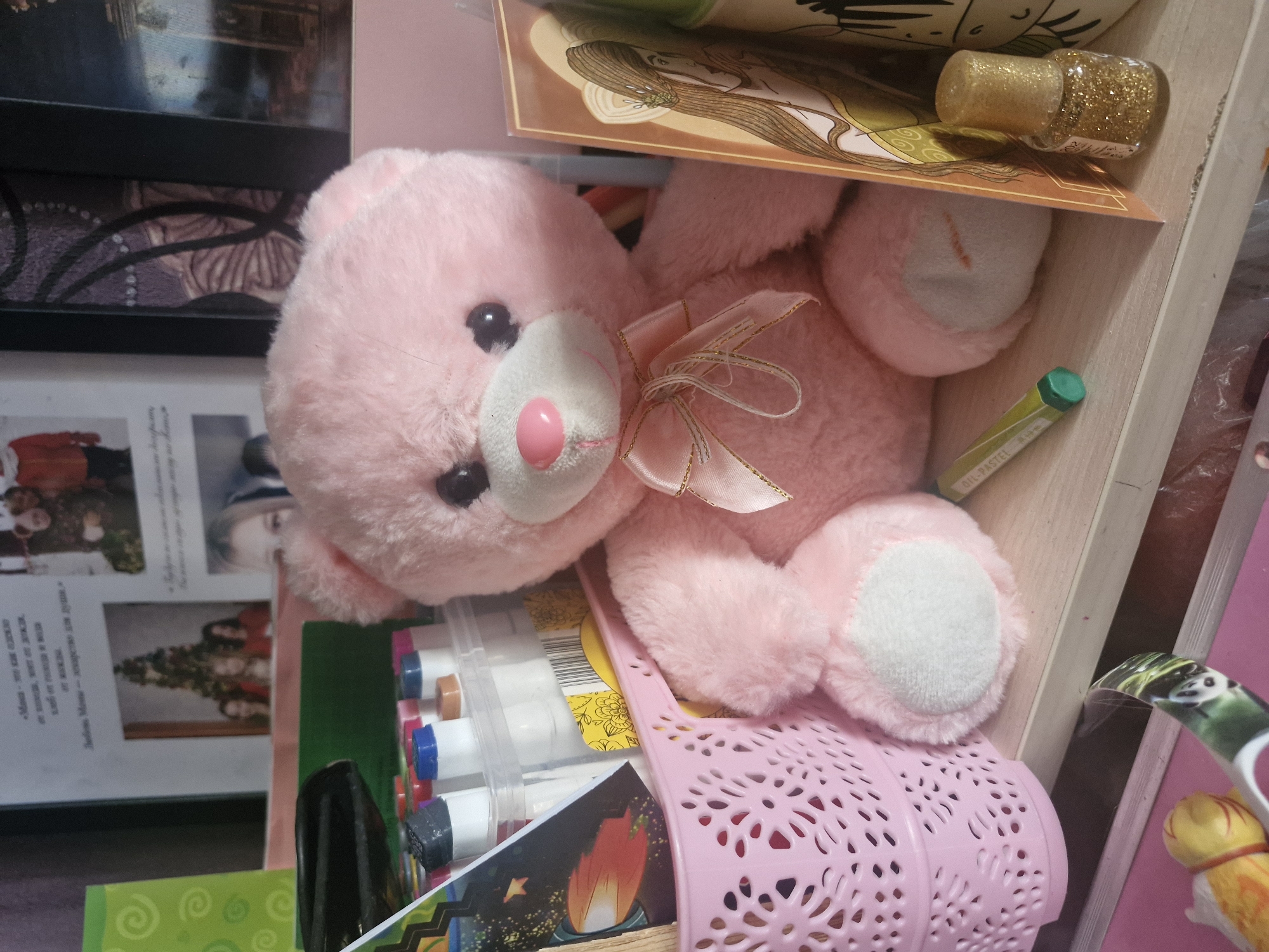 The image shows a close-up of various objects. Predominantly visible is a pink teddy bear with a white patch on its belly and feet. The bear has a pink bow tied around its neck and sits inside a pink, perforated plastic basket. The basket also contains a plastic case of markers and a card with an illustration of a hot air balloon. Above the teddy bear, there's a card with an illustration of a woman with long hair and a small jar of gold glitter. To the left, a picture frame displays photos and text. Other items visible in the background include a green crayon, a dog toy and additional papers and items on a shelf. The location of the image is unspecified.