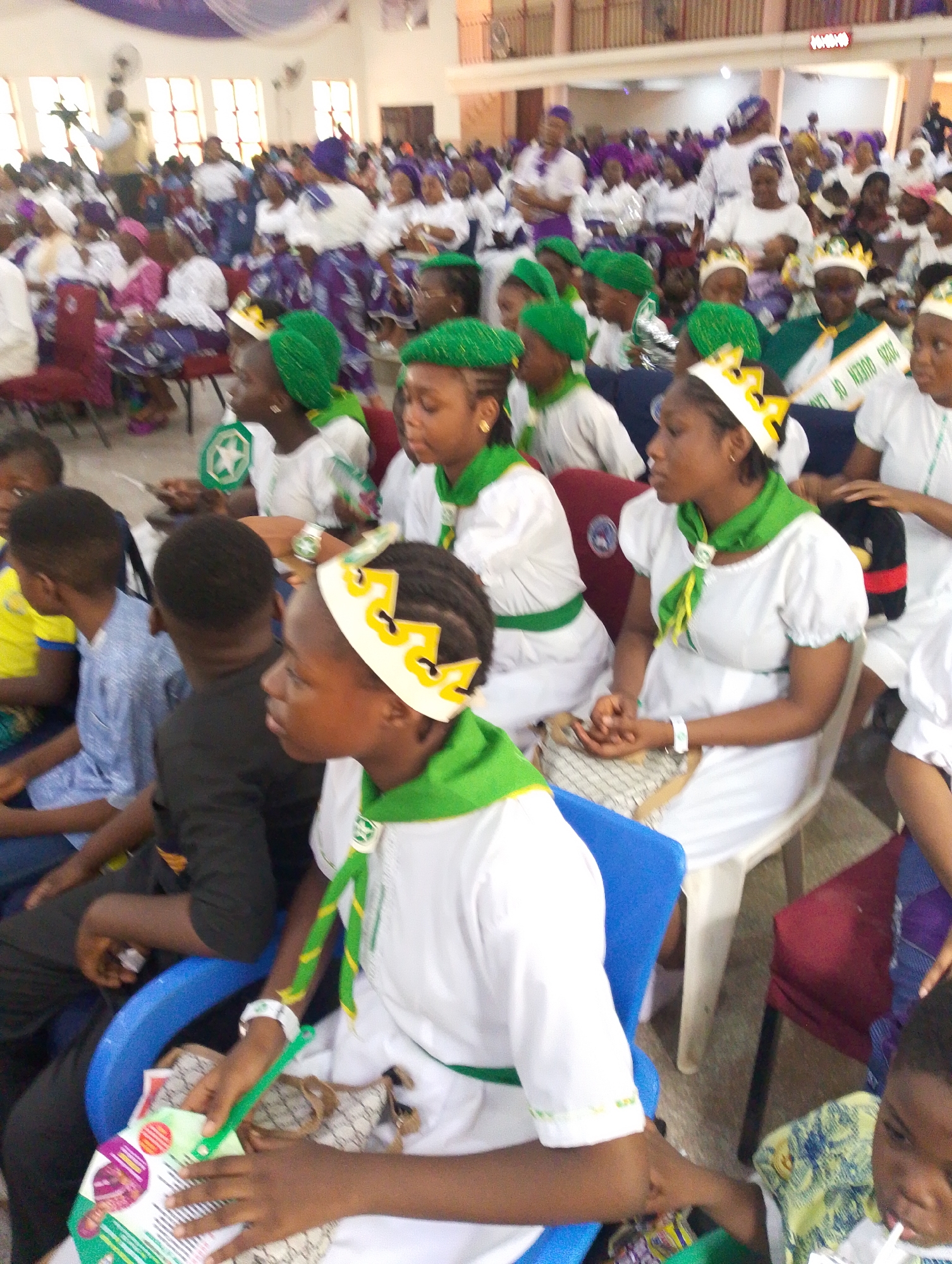 The image depicts a large gathering of people inside a building, possibly a church or event hall, in Kubwa, Nigeria. The crowd is predominantly comprised of women and children, many of whom are dressed in white garments with green accents, including head coverings, scarves, and belts. Some wear crown-shaped headbands. The people are seated in rows of chairs, suggesting an organized event or service. Some individuals hold green and white Nigerian flags or printed materials. The environment appears to be brightly lit, with white walls and a high ceiling.