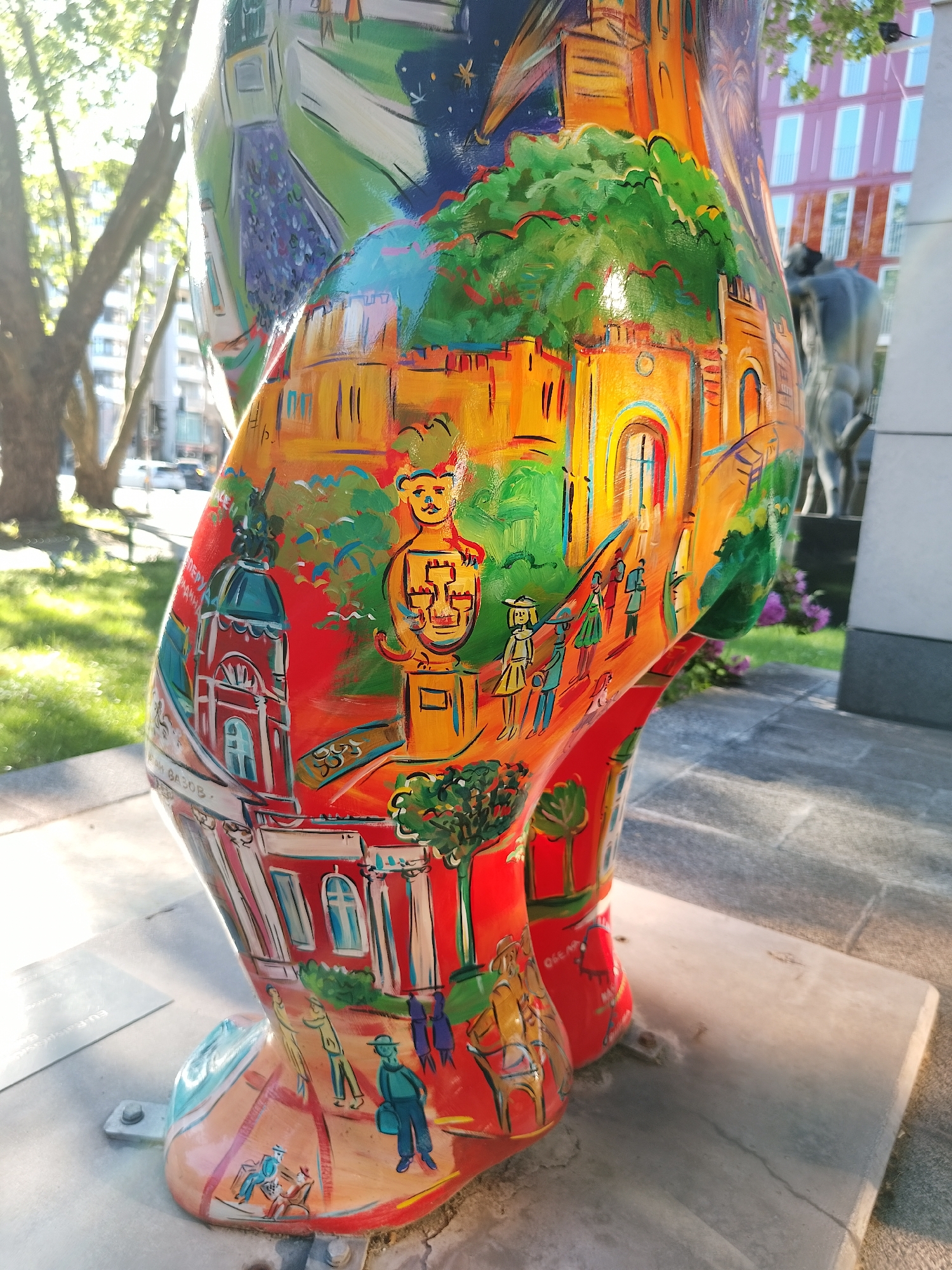 A vibrantly painted bear statue stands on a concrete base in an outdoor city setting. The bear's body is covered in a colorful mural depicting various scenes, including building facades, figures walking, a domed structure, and a cartoonish bear figure. The colors used in the mural are primarily reds, yellows, greens, and blues. A plaque is visible on the base near the bear's foot. In the background, there are trees, a street with vehicles, a building, and another statue. The light suggests it is daytime with sunny conditions. The location appears to be an urban park or public space.