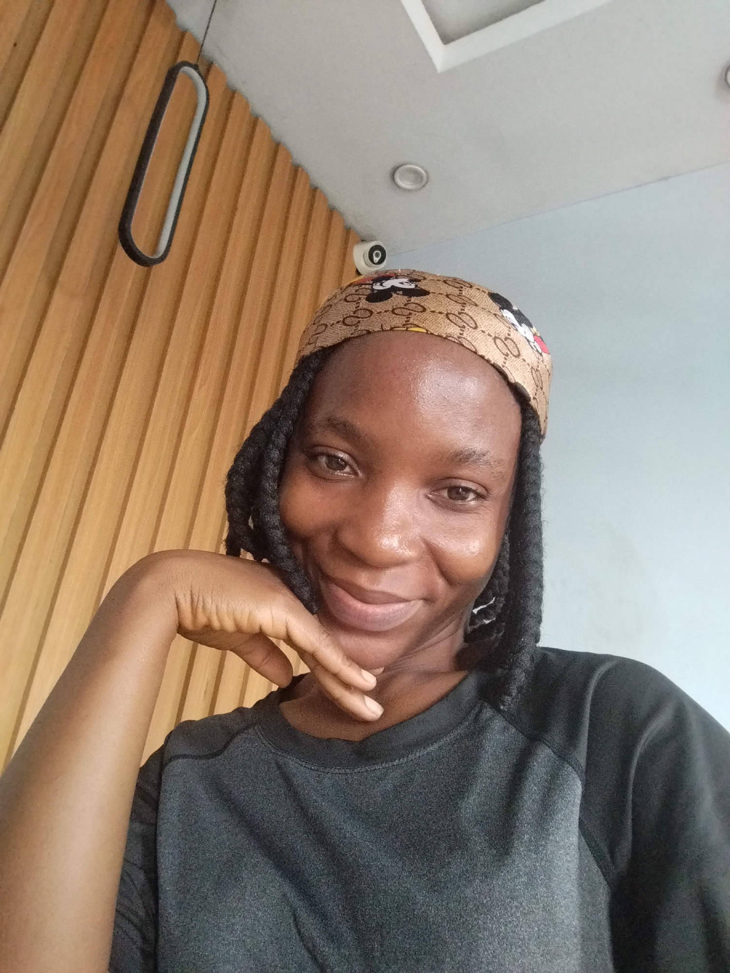 A young Black woman with braided hair and a headband is captured in a selfie. She is wearing a dark gray long-sleeved shirt and rests her chin on her hand, looking towards the camera with a slight smile. The background features a wall with vertical wooden paneling on the left and a light gray wall on the right. Above, a white ceiling with recessed lighting and a unique oval-shaped light fixture is visible. The overall scene appears to be indoors, possibly in a modern cafe or office setting.