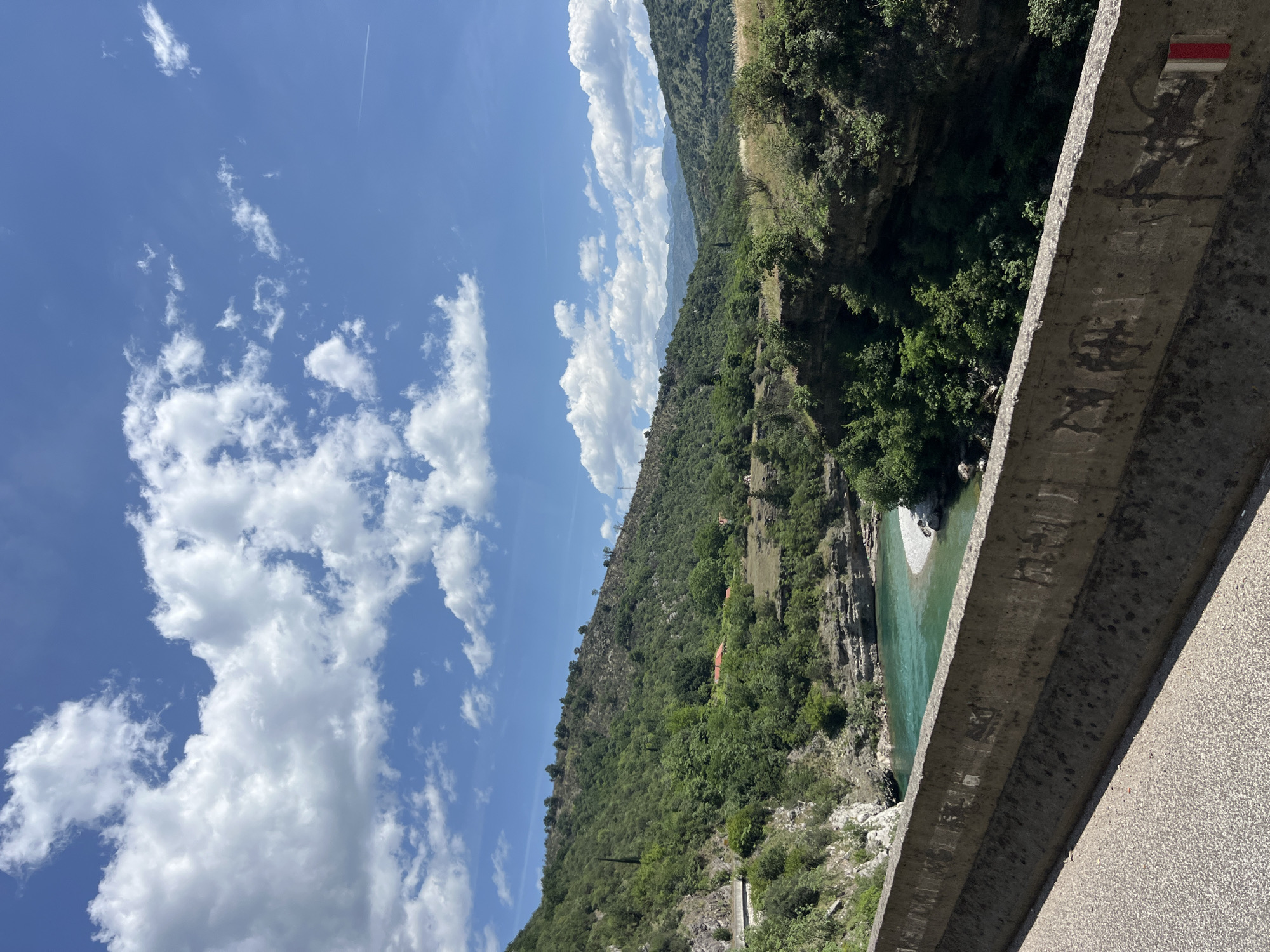 The image depicts a view from a bridge in Donje Mrke, Montenegro. The sky is blue with white, puffy clouds. Below the bridge, a turquoise river flows through a forested valley. The bridge's concrete railing is visible in the foreground. A red and white trail marker is affixed to the railing. Vegetation covers the steep hillsides surrounding the river.