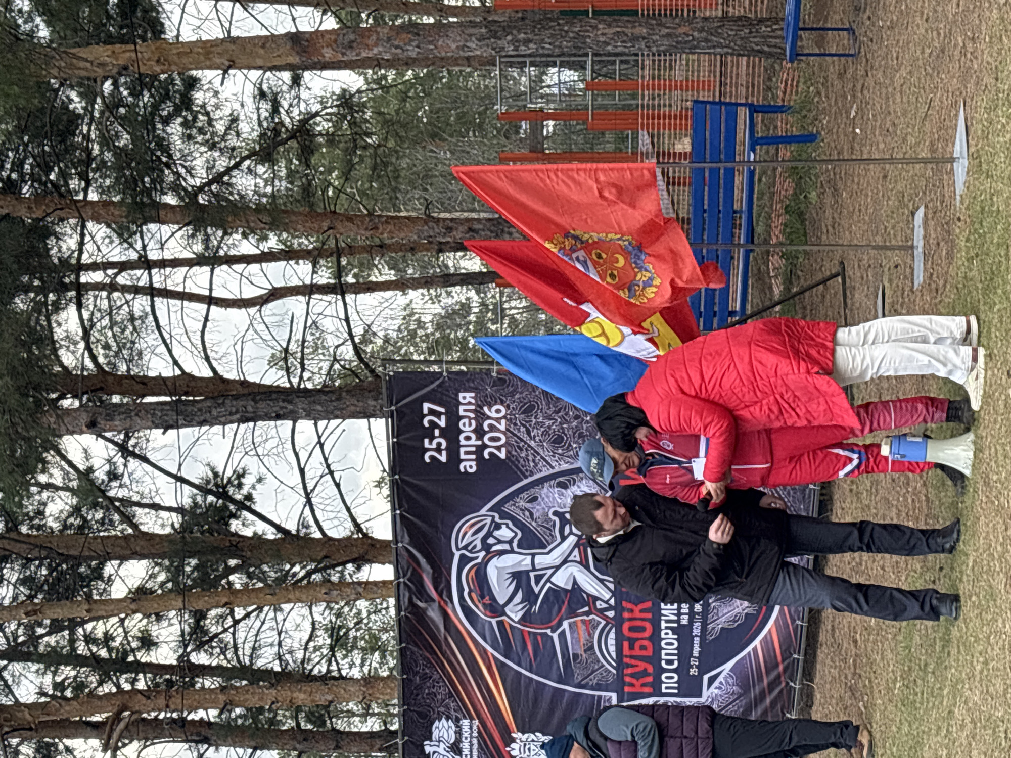 This media file, captured in Orenburg, Russia, depicts an outdoor scene set up for a sporting event, likely a cycling competition given the graphic on the main banner. The prominent banner, with a black background, features a white illustration of a cyclist and advertises the "SPORTS CUP" (КУБОК ПО СПОРТИЕ) scheduled for April 25-27, 2026. The text on the banner also includes "г. ОРЕНБУРГ" (Orenburg), confirming the event's location.
Several individuals are present. Two people are dressed in athletic wear, one in a red jacket and white pants, and another in a full red tracksuit, appearing to be engaged in a discussion or preparation. A man in a black jacket stands next to the banner, observing. Other figures are partially visible in the foreground and background.
The setting is outdoors, characterized by numerous tall pine trees under an overcast sky, suggesting daylight without direct sun. Blue park benches and metal fencing are visible in the background, indicative of an organized event space. Multiple flags are flying, including a distinct red flag adorned with a coat of arms featuring a yellow shield, a red cross, and three fleurs-de-lis, possibly representing a sponsoring region or organization. A blue megaphone rests on the ground near the participants, hinting at event coordination or announcements. The image captures a moment during the event or its setup, conveying an atmosphere of sporting activity.